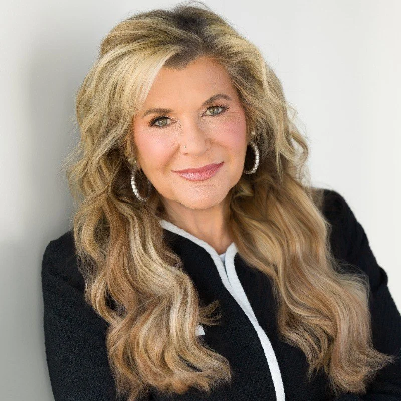 A woman with long, wavy blonde hair and light skin, wearing a black top with white trim and large hoop earrings, smiling slightly at the camera.