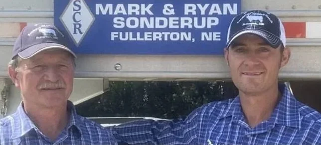 Two men in blue plaid shirts standing in front of a sign that reads "Mark & Ryan Sonderup Fullerton, NE"