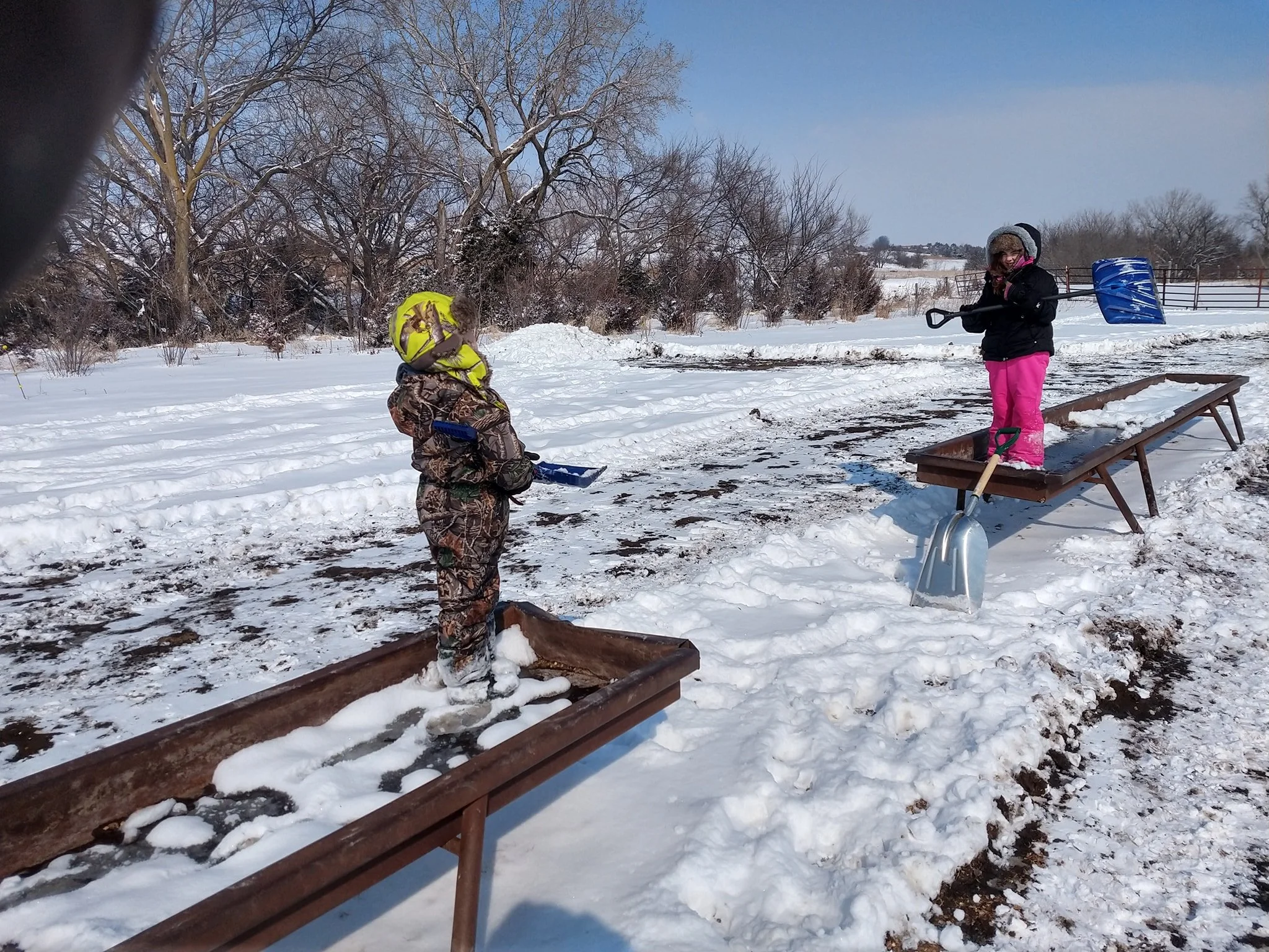 Two children in winter clothing playing outdoors in the snow, standing on rails and a sled with snow shovels in hand, surrounded by trees and open fields.