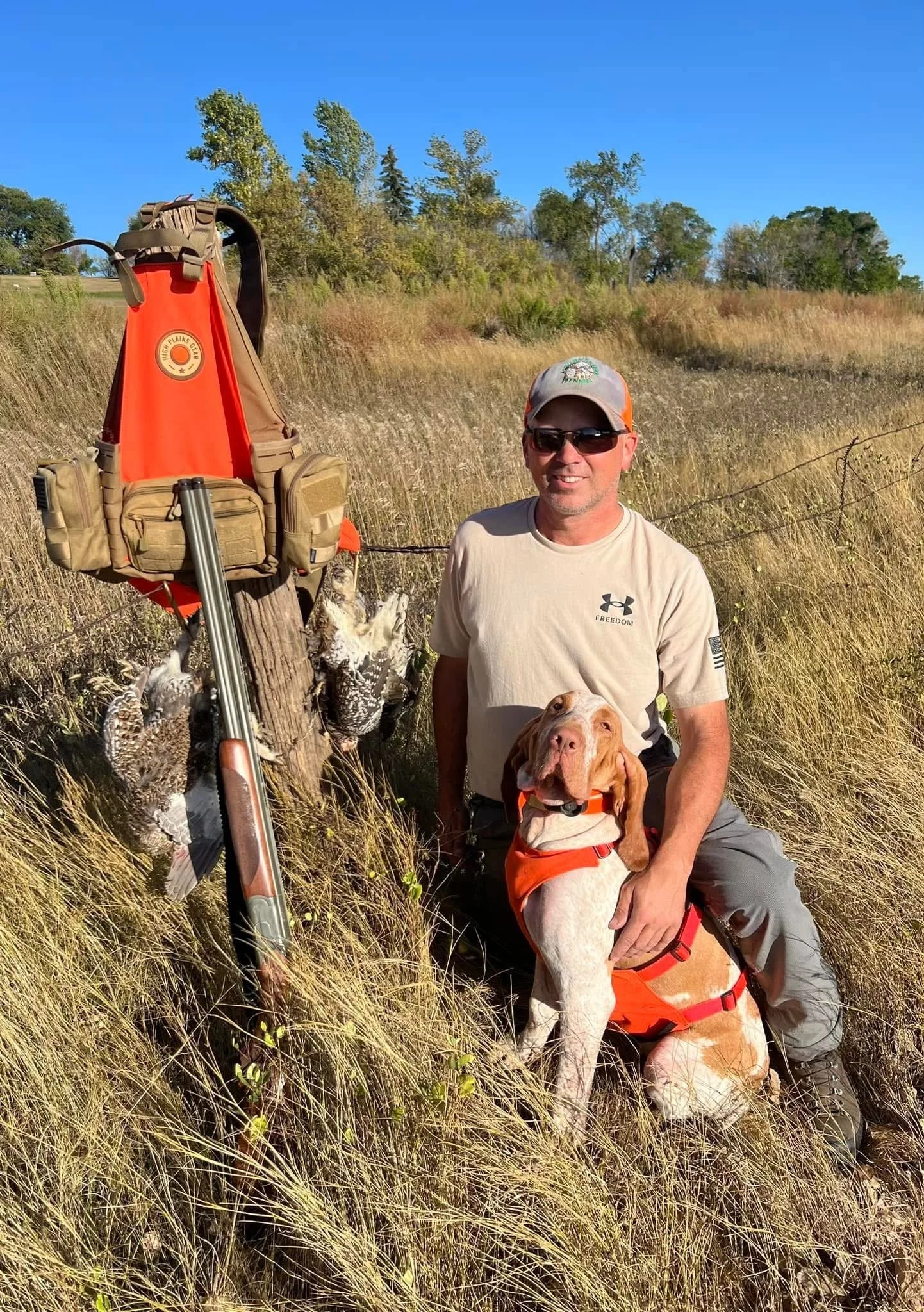 A man in sunglasses and a beige t-shirt kneels next to a hunting dog in an orange vest, beside a tree with hanging hunting decoys and a rifle, in a grassy field under a clear blue sky.