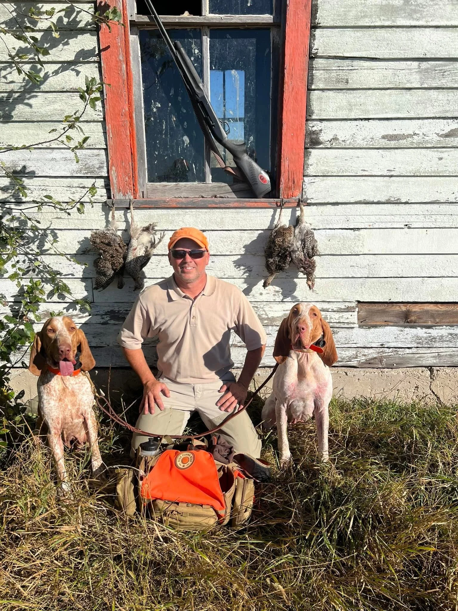A man with two hunting dogs and two dead birds hanging on the wall outside an old, weathered wooden building with a window, a shotgun leaning against the window, and hunting gear on the ground.