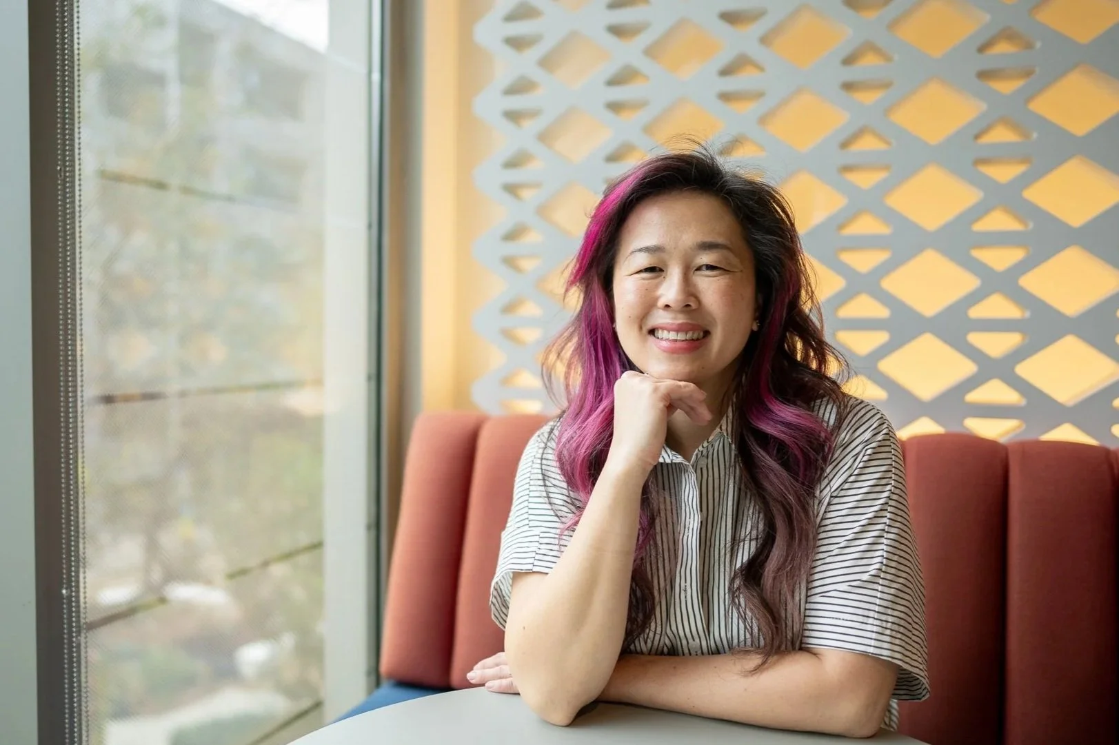 Women with pink purple hair sitting in a pink color booth with her elbow on the table while resting her head on the back of her fingers.