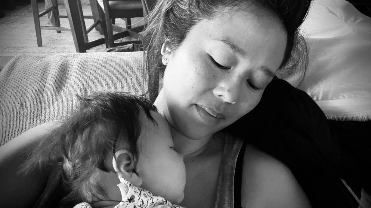 Woman sitting in a couch with her eyes closed while her baby sleeps in her arms.