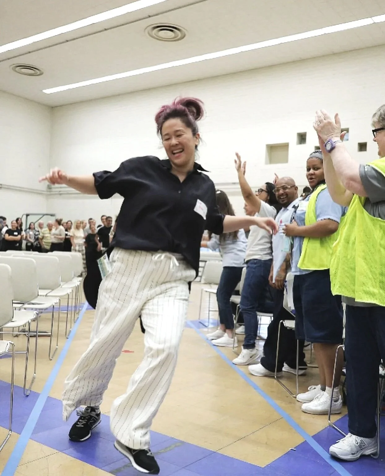 Woman with pink hair dancing down the aisle with her arms outstretched in hip hop style; moving away from the crowd of volunteers.