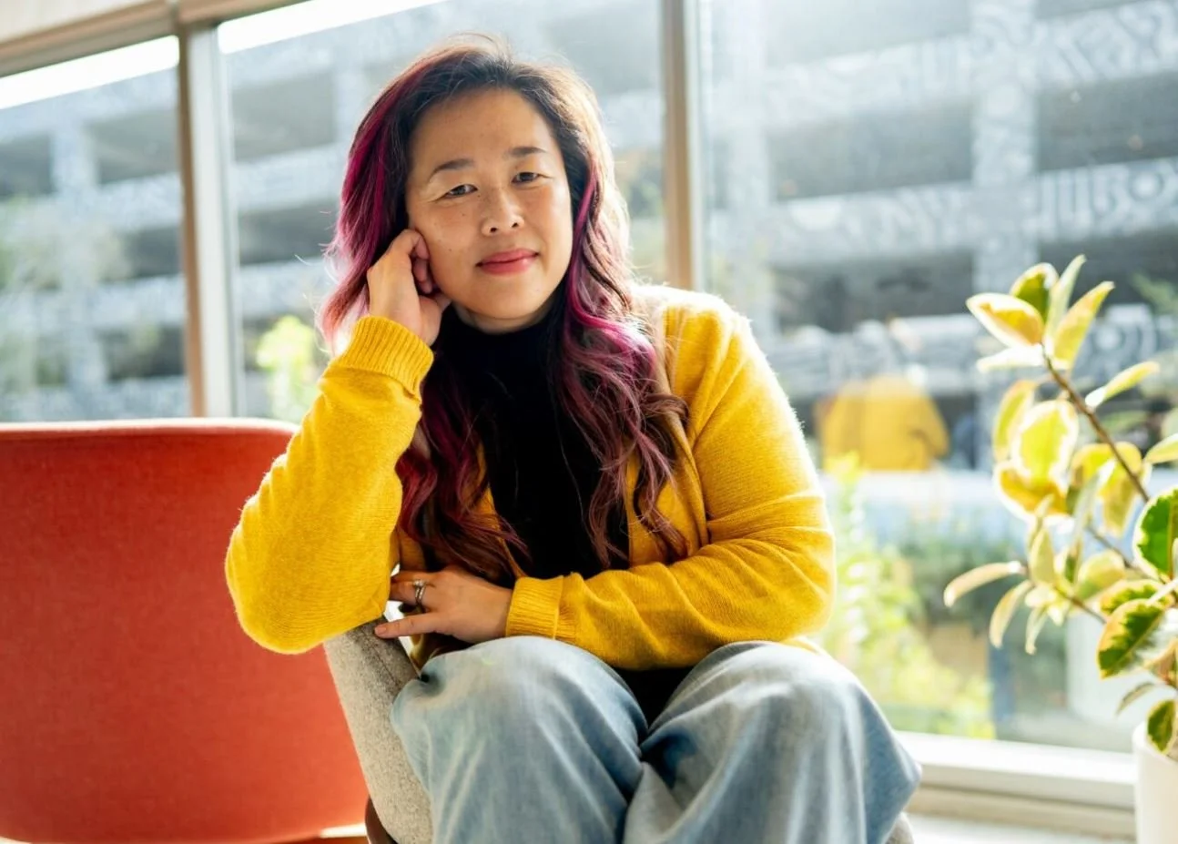young asian women with pink highlights sitting in a small chair with elbow propped on the back of the chair with her head resting against her fingers in a gentle fist wearing yellow cardigan and light blue jeans