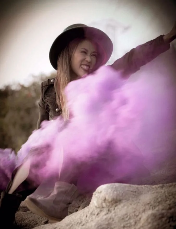 Woman in dark attire wearing a medium size brimmed hat pointing behind an explosion of pink and purple cloud of smoke