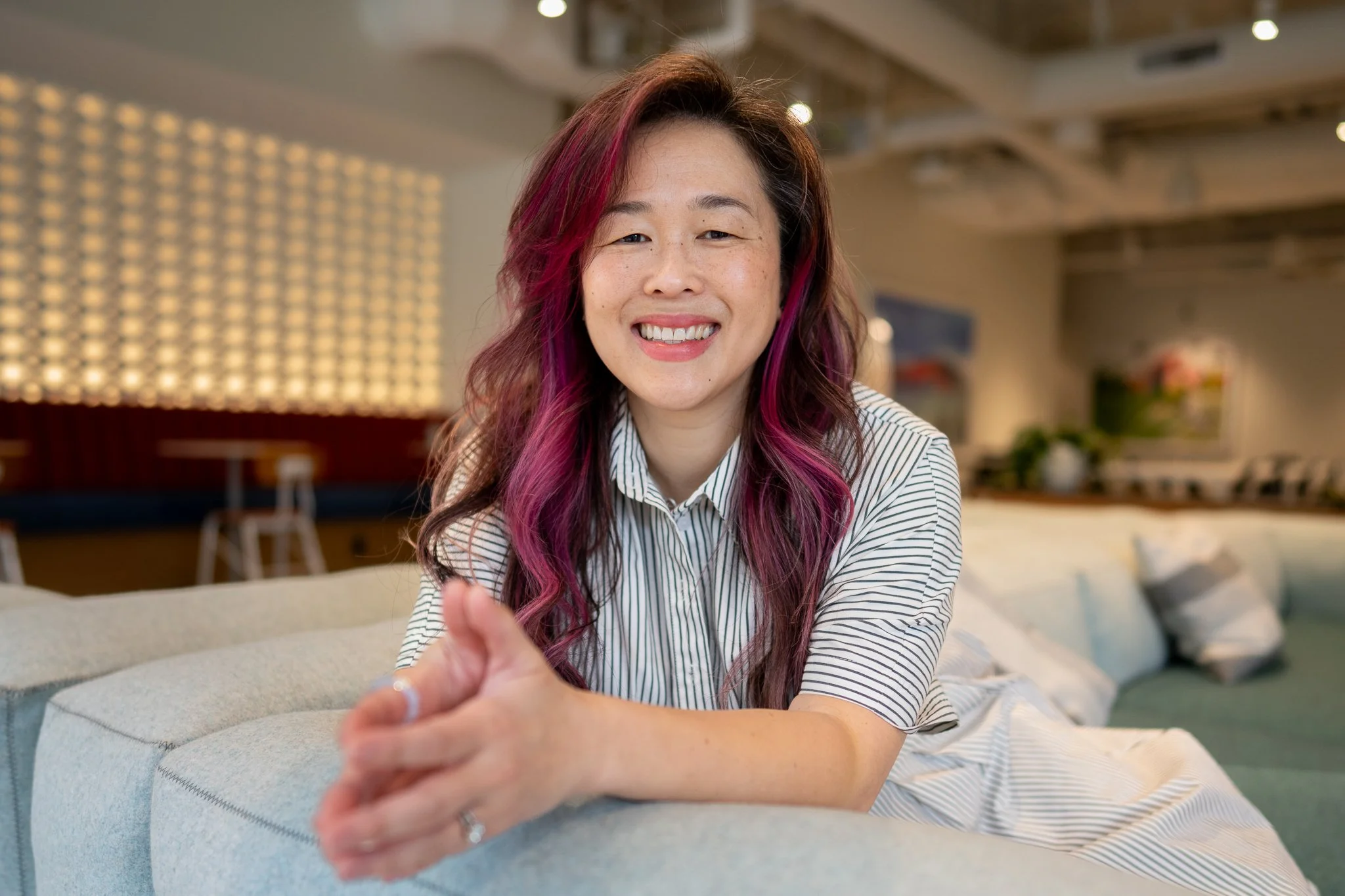 Woman with pink highlights and auburn hair sitting on the couch smiling with her palms pressed together; wearing a casual short sleeve button shirt inside a modern comfy activity room.