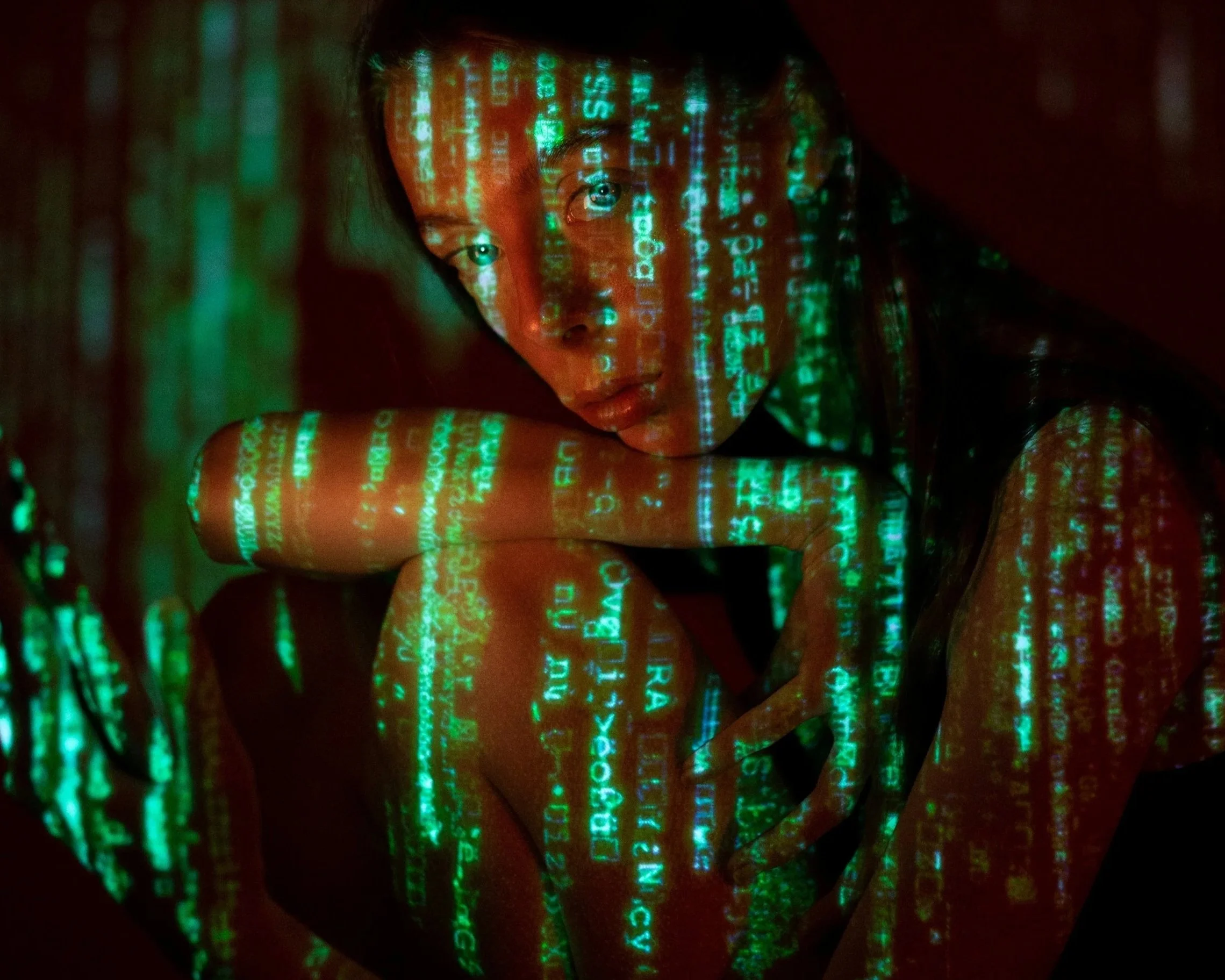 Young woman sitting in the dark with her arm and chin resting on her knee, neon bright letters cover her face and body. Cyberpunk and matrix vibe.