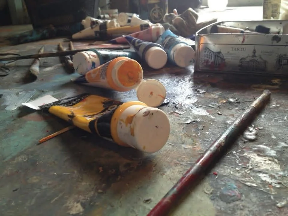 Numerous used bottles of acrylic paint and paintbrushes with paint splatter on top of a very large dark color painted canvas with a white box of paint in the background.