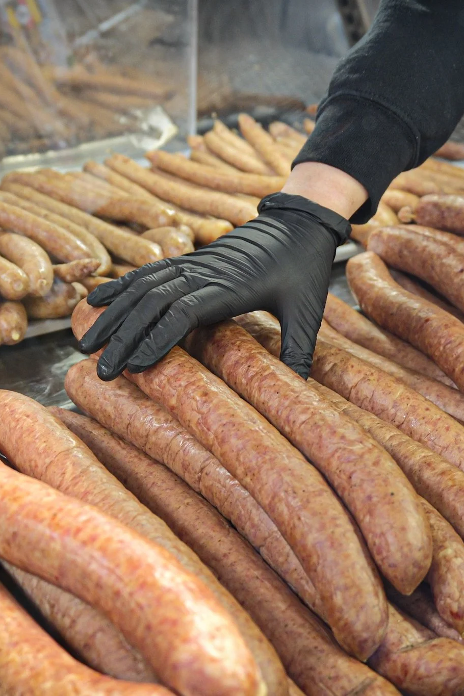 A person wearing a black glove is handling raw sausages at a sausage-making or butcher shop, with multiple raw sausages laid out on a metal surface.