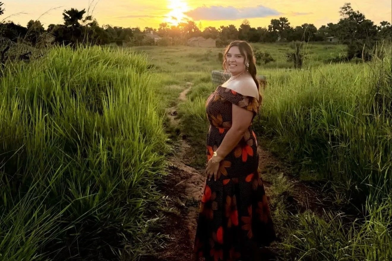 A woman in an off-shoulder dress with a floral pattern standing on a dirt path through green fields at sunset.