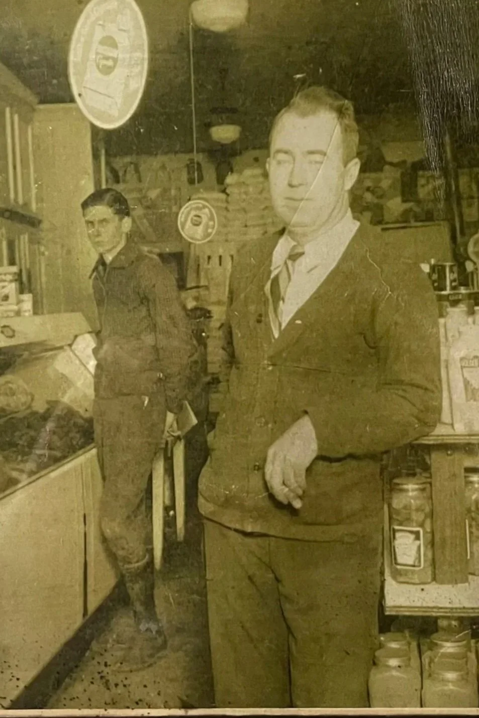 A vintage black and white photo showing two men in a store. One man stands in the foreground with a serious expression, dressed in a suit and tie, with his arm resting on the counter. The other man, in the background, is also dressed in a suit, holding a newspaper, and appears to be looking elsewhere.