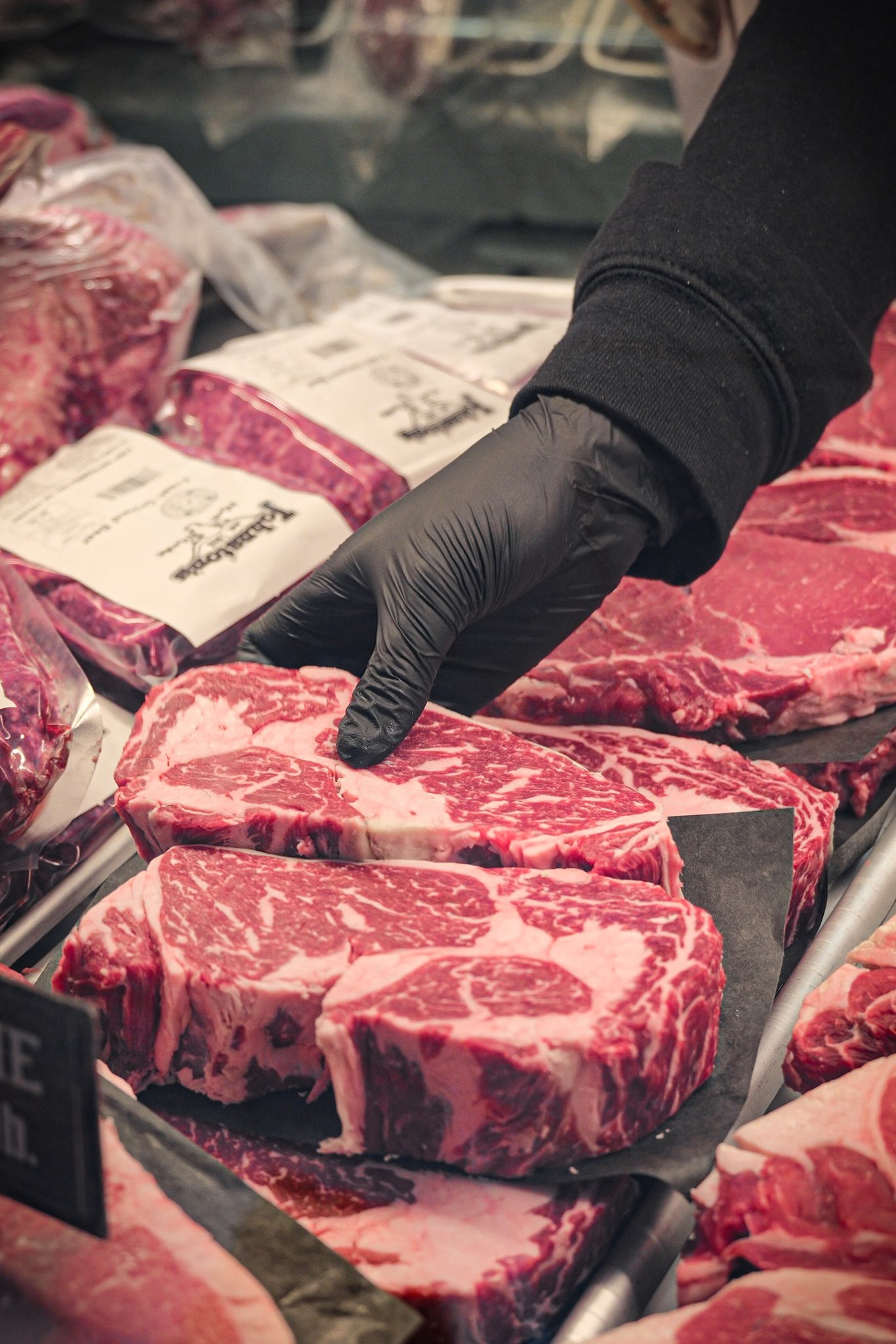 A person wearing a black sleeve and black gloves handling several raw, marbled beef steaks at a butcher shop or meat market.