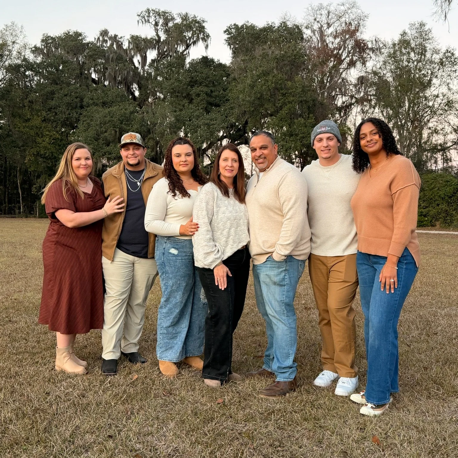 Group of eight people standing outdoors on grass with trees in the background, smiling and posing for a photo.