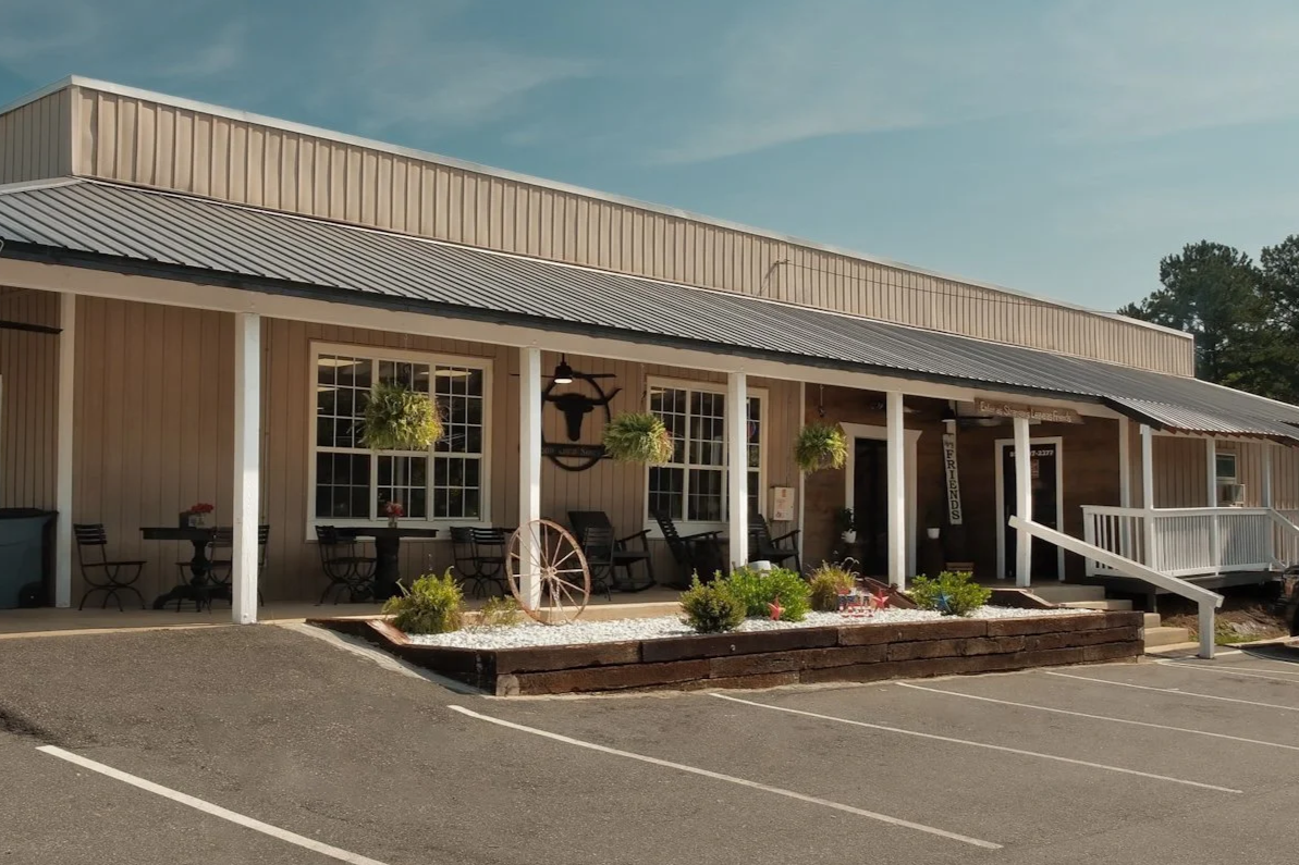 A beige building with a metal roof, outdoor seating, hanging plants, and decorative items outside, including a wagon wheel and a bull's head sign, in front of a parking lot.