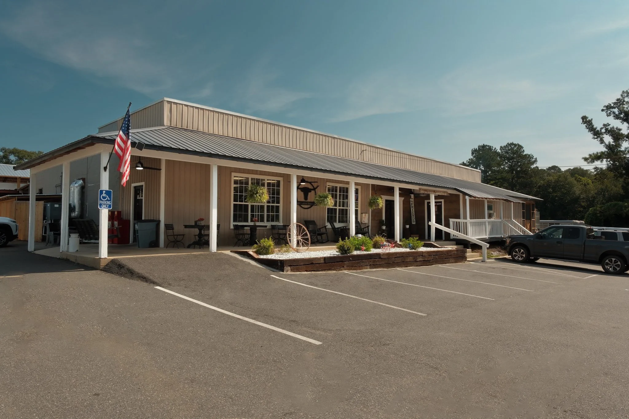 A small beige commercial building with a metal roof, outdoor seating, and hanging plants, located in a parking lot with marked spaces and a handicapped parking sign, under a clear blue sky.