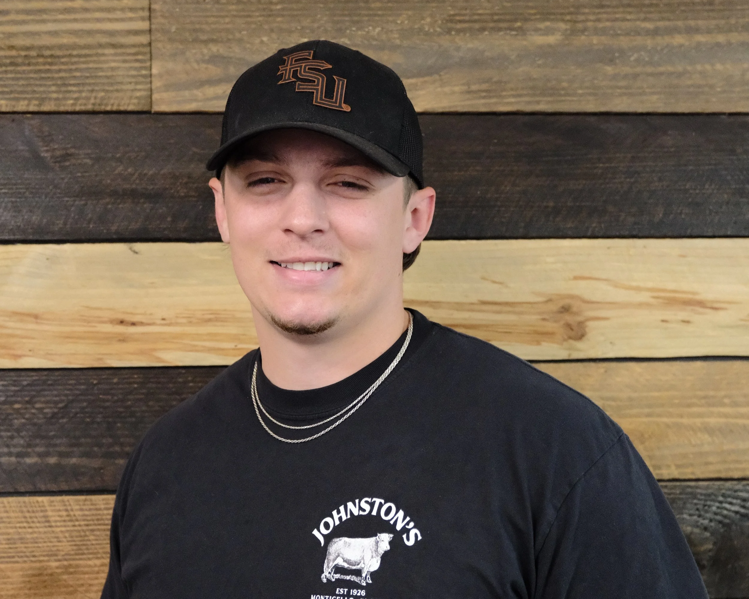 Close-up of a young man wearing a black baseball cap with orange embroidery and a black T-shirt with white graphic and text, standing against a wooden wall background.