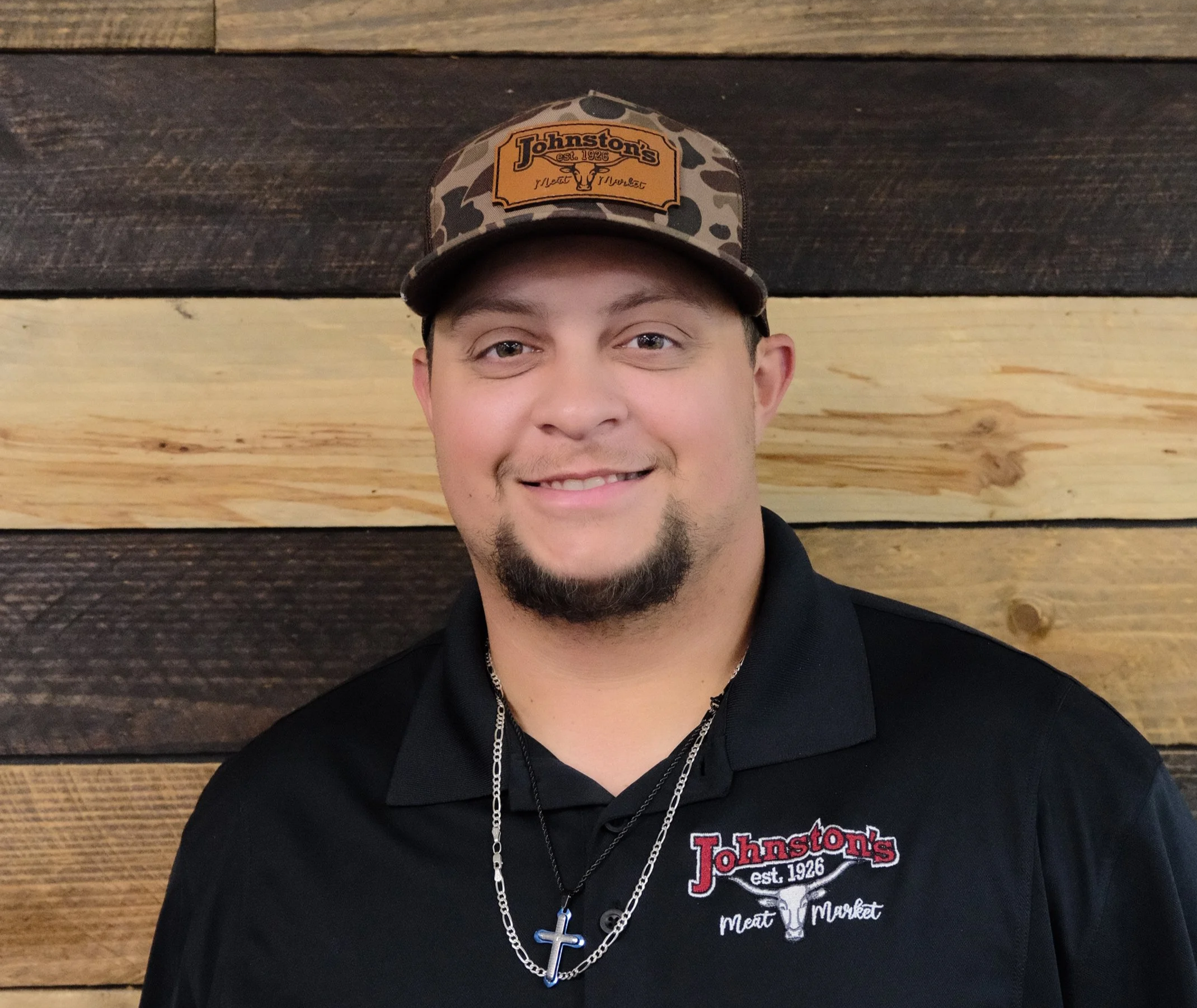 A young man with light skin, short dark hair, and facial hair, smiling, wearing a camouflage baseball cap with a 'Johnston's' logo and a black polo shirt with a 'Johnston's' logo, standing in front of a wooden wall with horizontal planks.