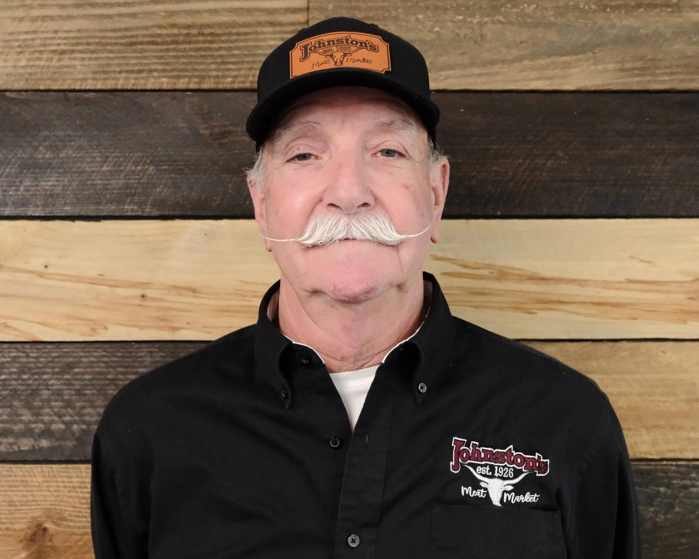 Close-up of a man wearing a black cap with an orange logo and a black shirt with a logo that says 'Johnston's Est. 1926 Meat Market' against a wooden plank background.
