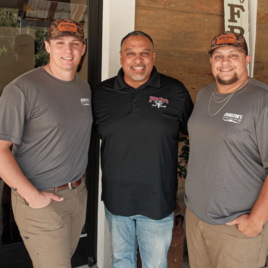 Three men smiling, standing outside a restaurant. The two younger men on either side are wearing gray Johnston's shirts and camo hats. The man in the middle is wearing a black shirt with Johnston's logo. They appear to be friends or colleagues.