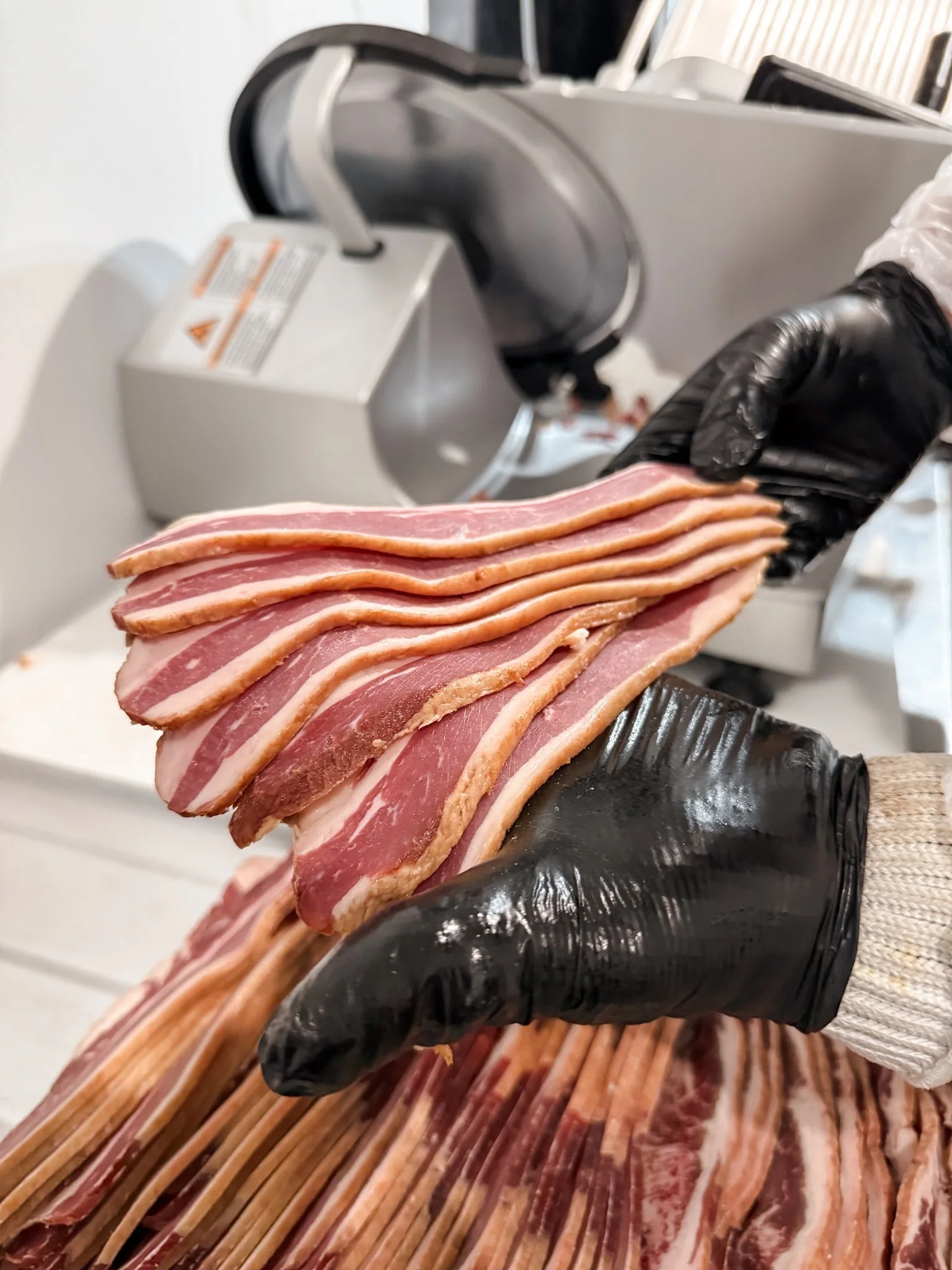 A person wearing a black glove is holding a stack of raw bacon strips in front of a meat slicer machine.