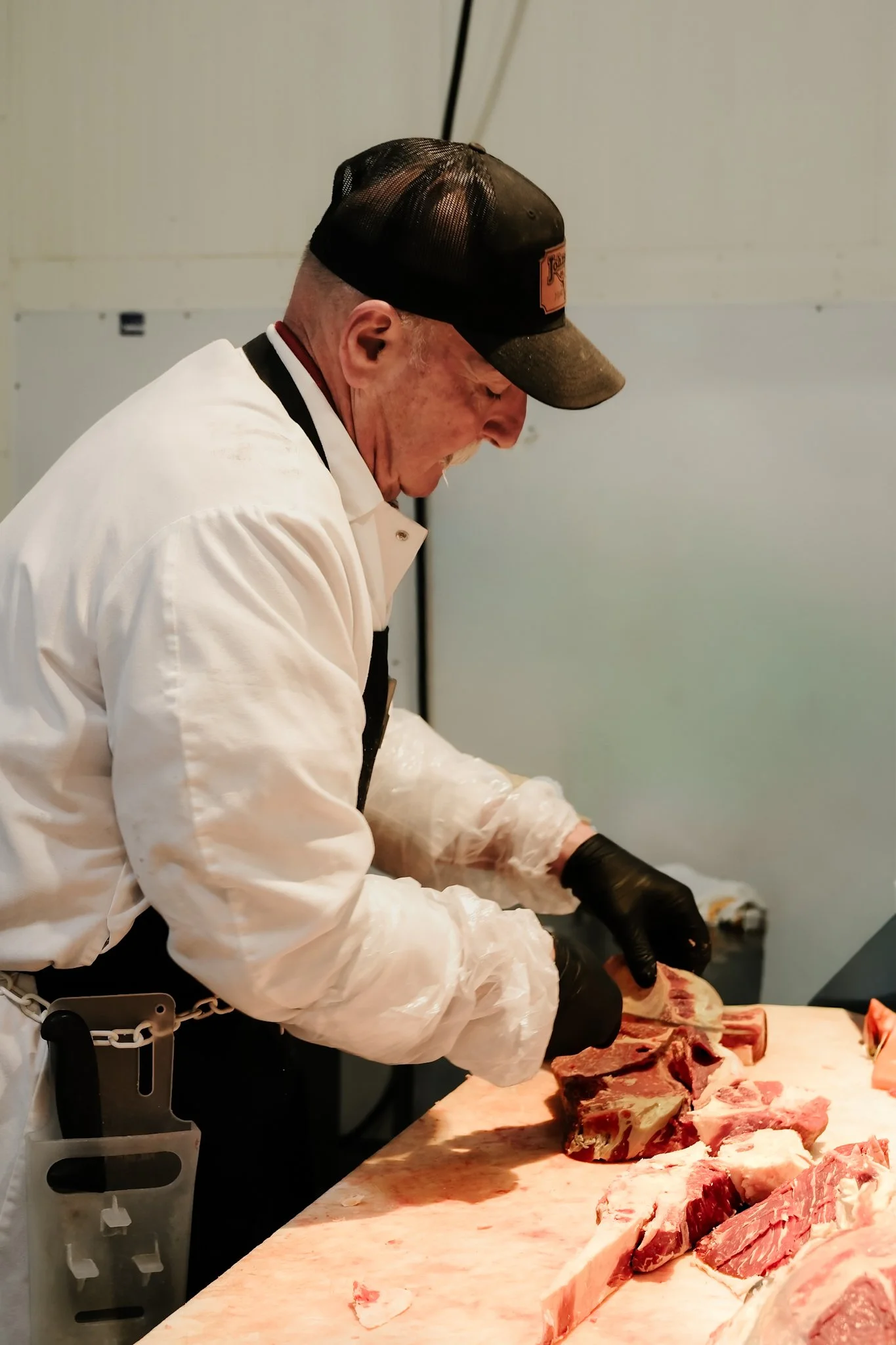 A man in a white coat and black gloves is slicing large pieces of raw beef at a butcher's station.