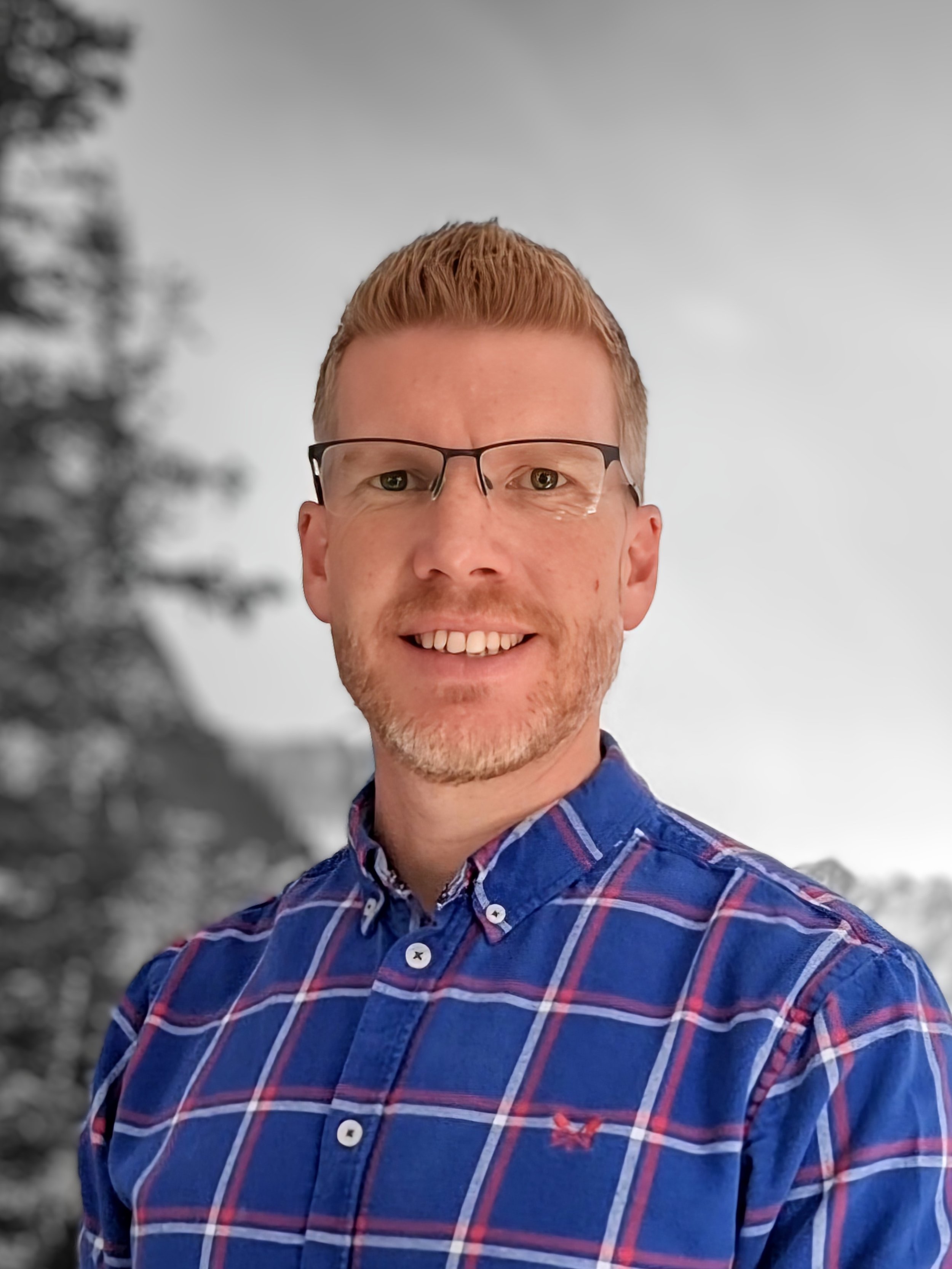 A man with short red hair, glasses, and a beard, smiling, wearing a blue plaid shirt, standing outdoors with blurred trees and sky in the background.