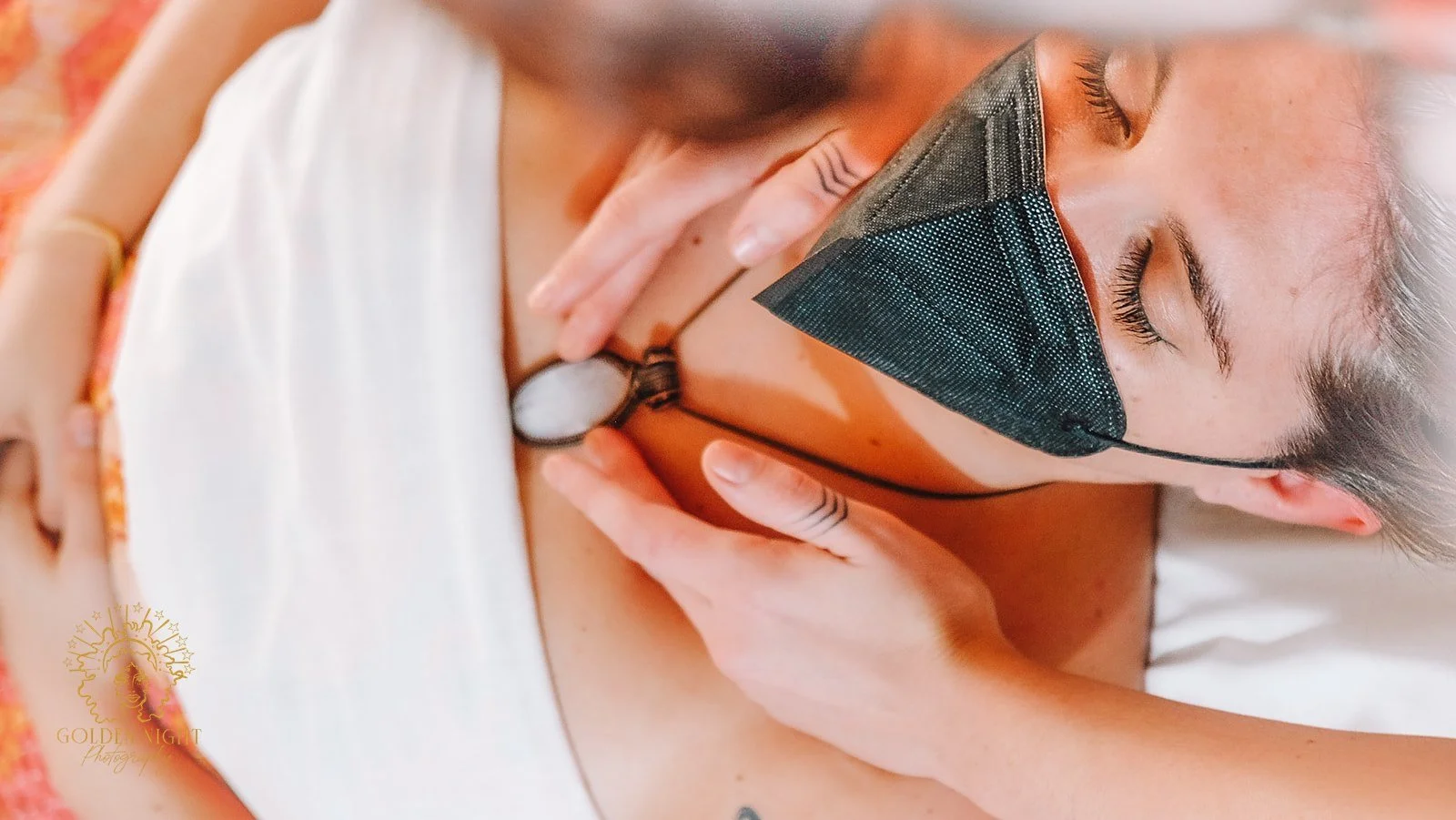 a white woman in a mask is massages at the collar bones around a necklance by white hands with finger tattoos