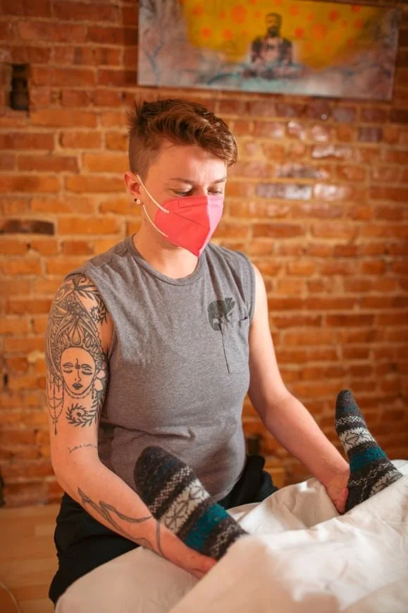 a white nonbinary person in a pink mask with red hair and tattoos cradles the ankles of a person wearing socks.