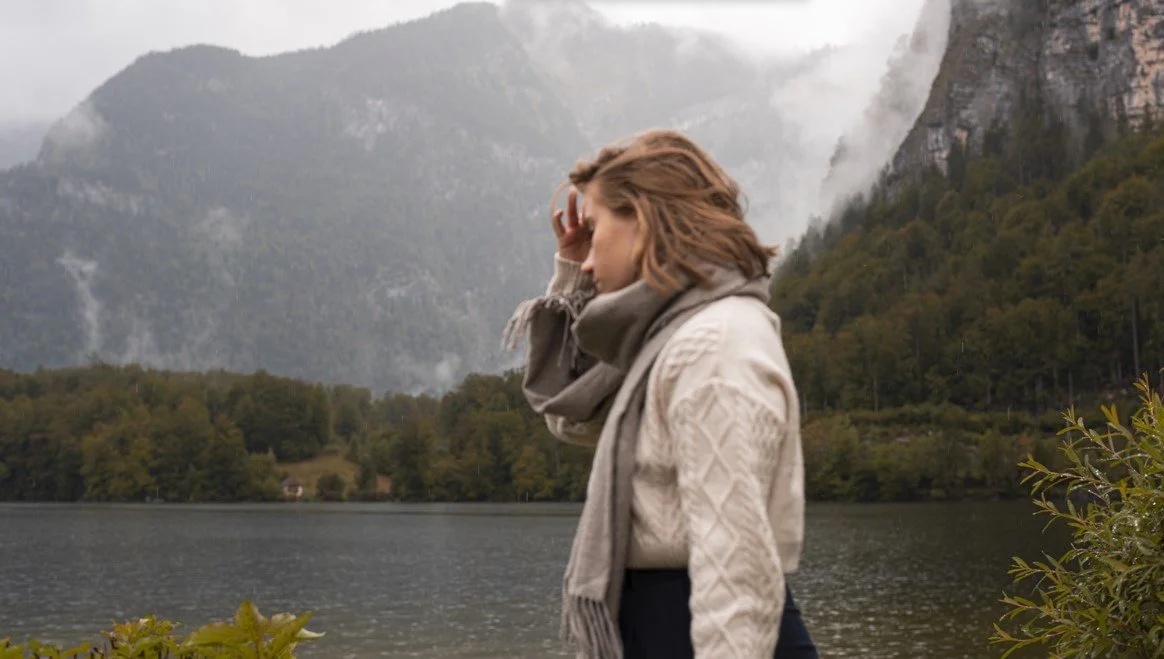 A woman standing near a lake with mountains and trees in the background, touching her face with her hand.