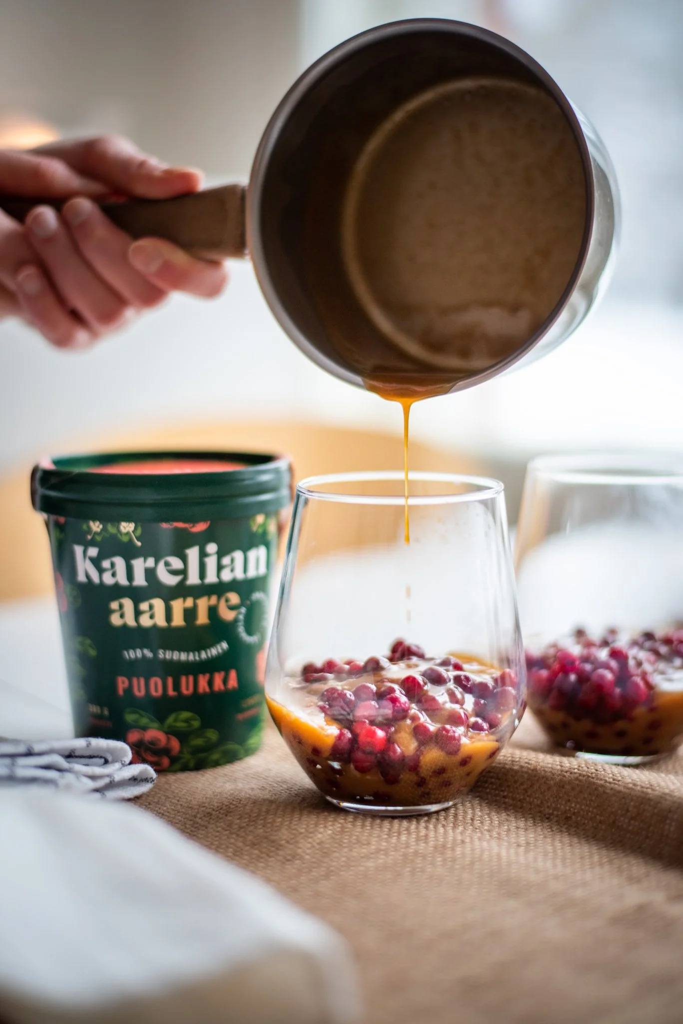 Pouring hot beverage into a glass with berries. A container labeled 'Karelian aarre' is in the background.
