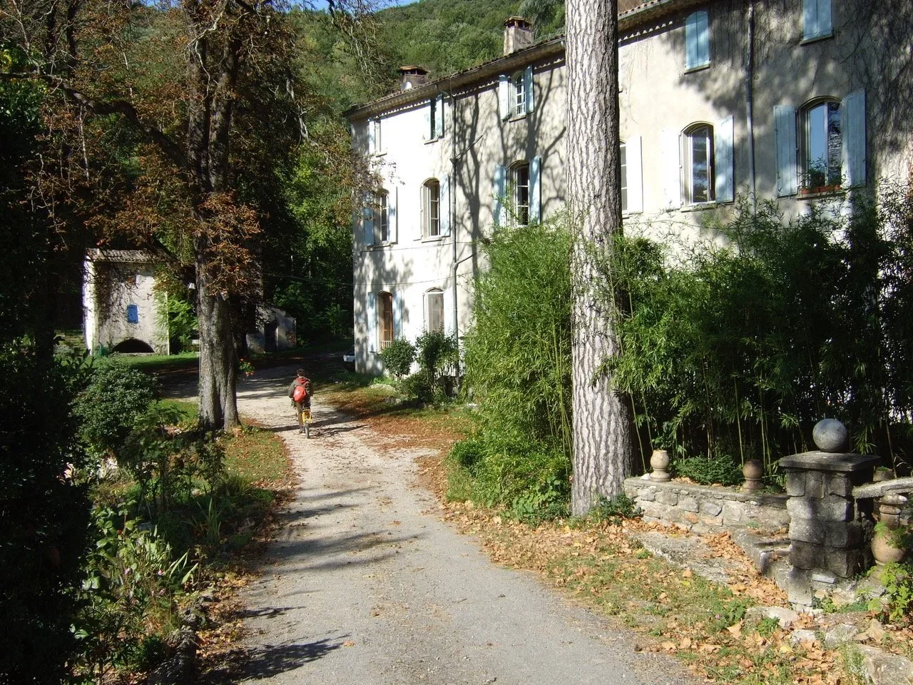 Front entrance view of the Château de Malérargues in the summer, a white building with multiple windows, in a scenic, wooded area, gravel path with a person riding a bicycle, surrounded by trees and greenery.