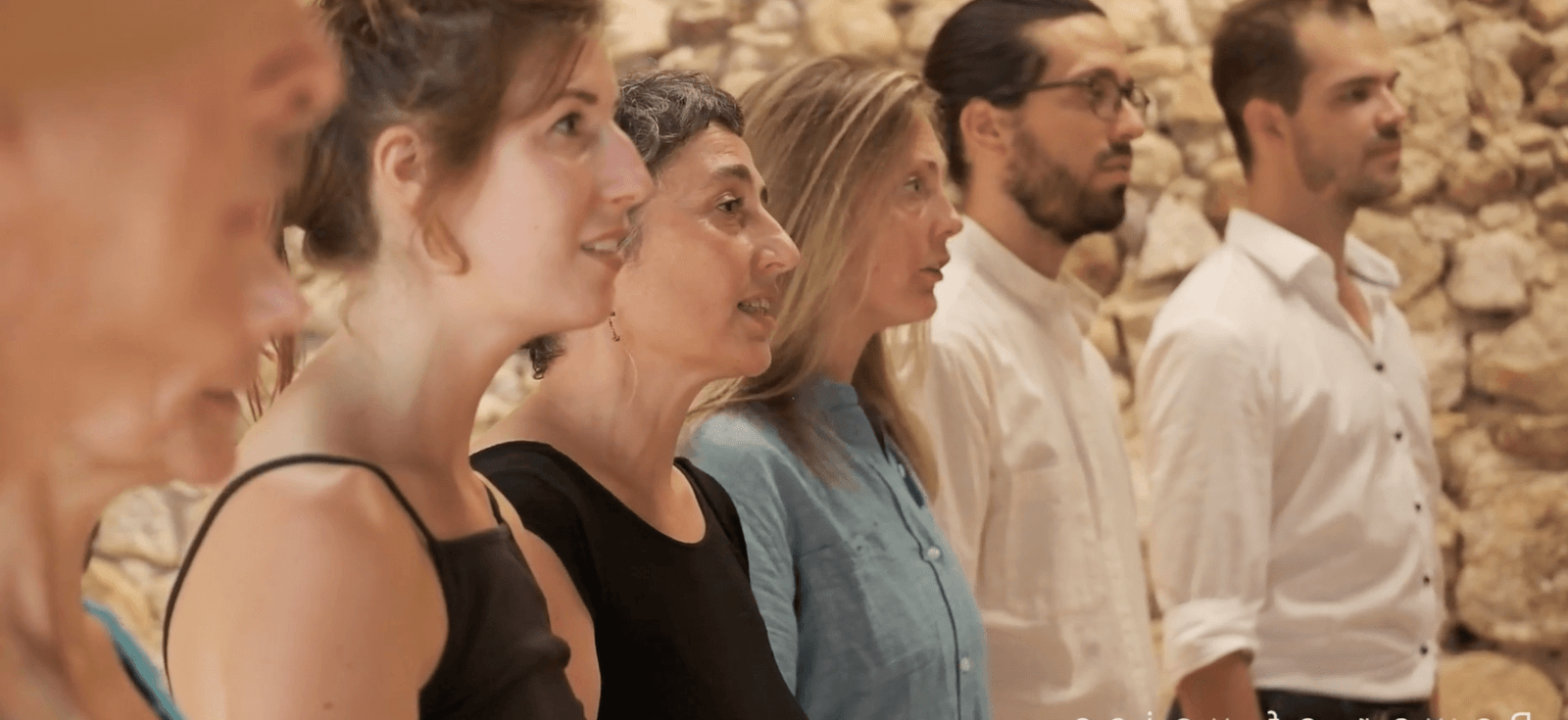 Six people standing side by side, engaged in a workshop activity, listening attentively, against the stone wall background of a studio in Malérargues.