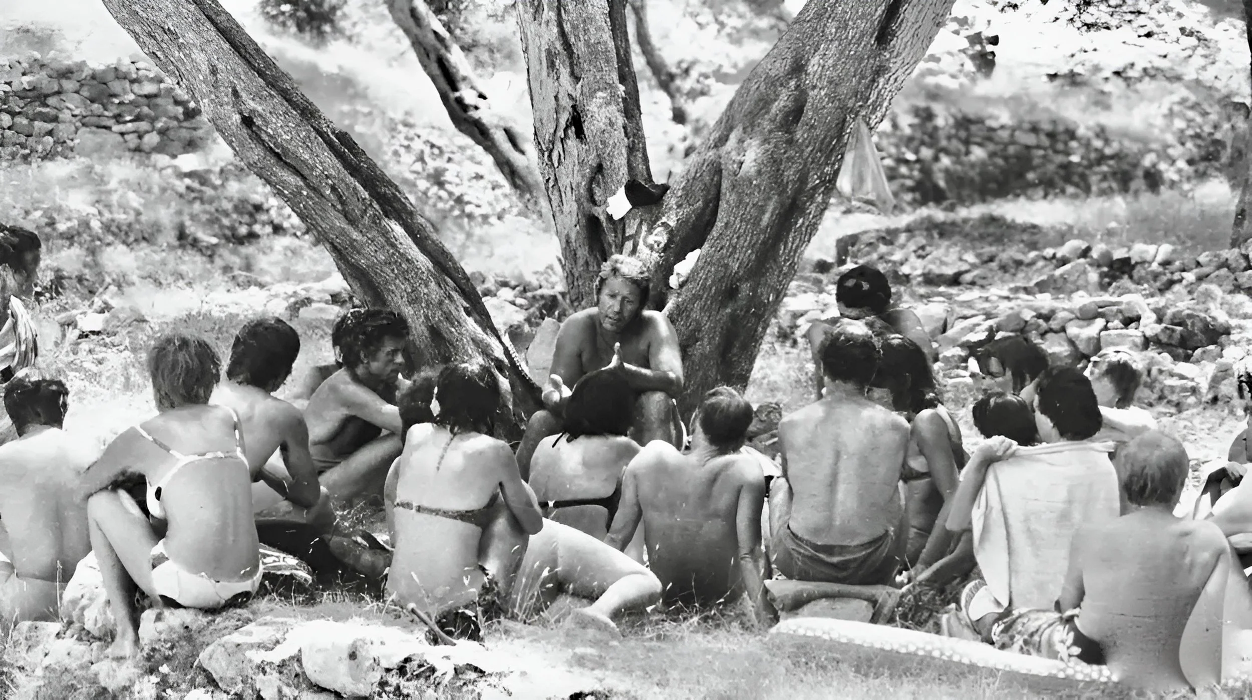 Black and white photo of members of the Roy Hart Theatre sitting outdoors in bathing suits near a large tree, engaging in a discussion with Roy Hart speaking while others listen. The setting is the island of Paxos in Greece in 1973.