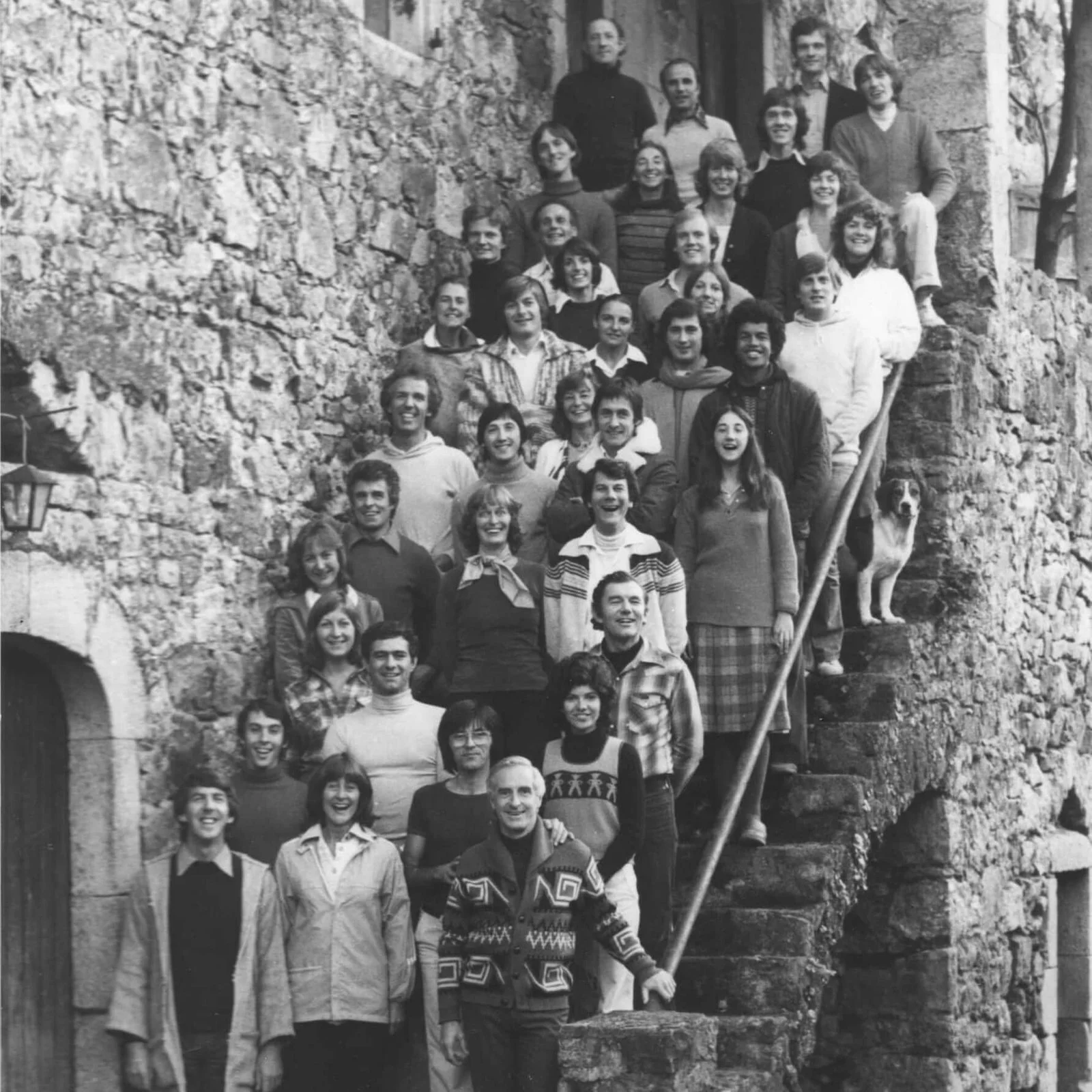 Members of the original 49 founders of the Roy Hart Theatre who moved to Malérargues, gathered on a stone staircase leading to the Magnanerie Theatre, outside, with a dog visible on the right side. The group is posing for the photo.