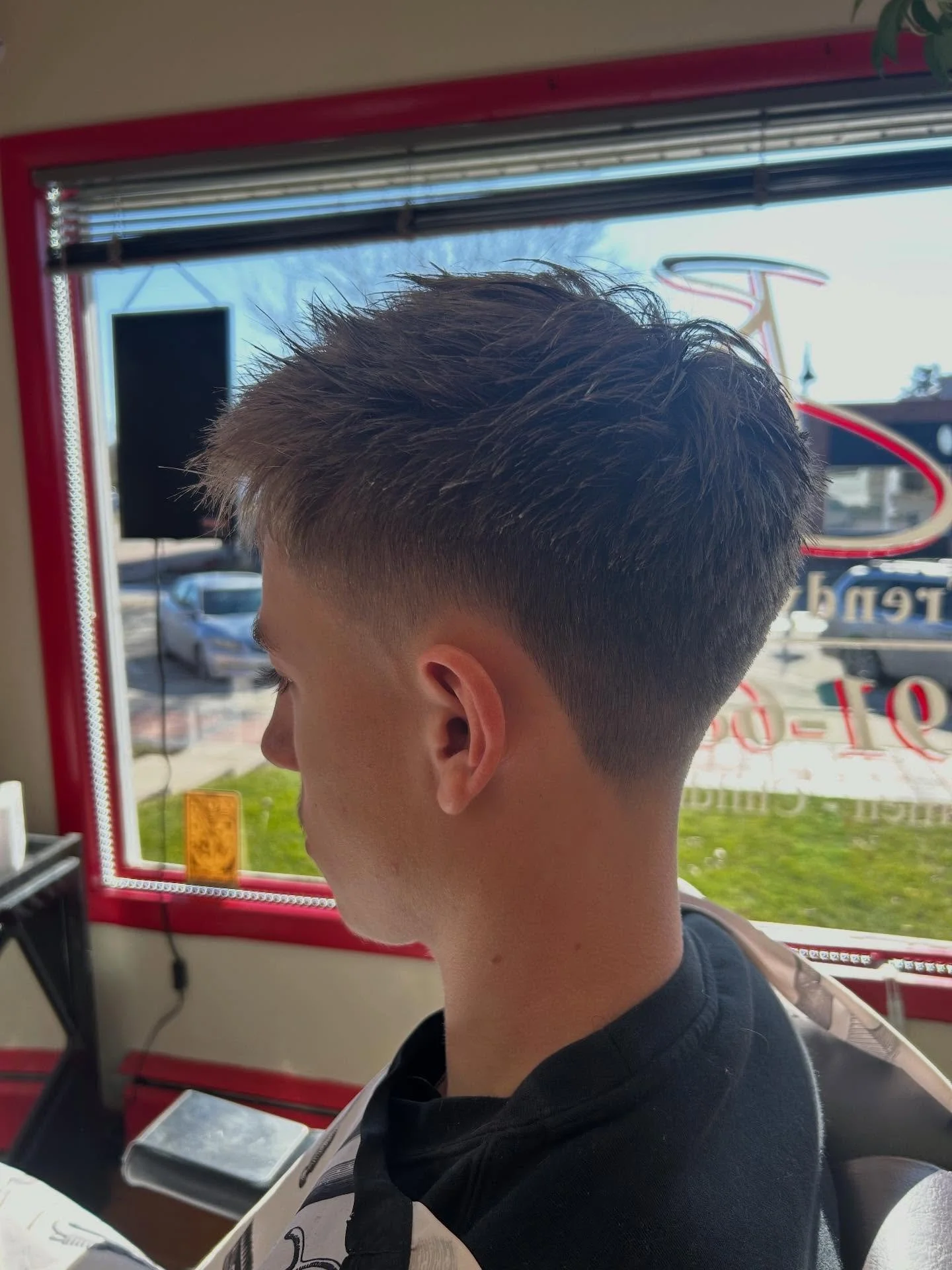 Side profile of a young man with a short, textured haircut sitting inside a vehicle, looking down at a device in front of him.