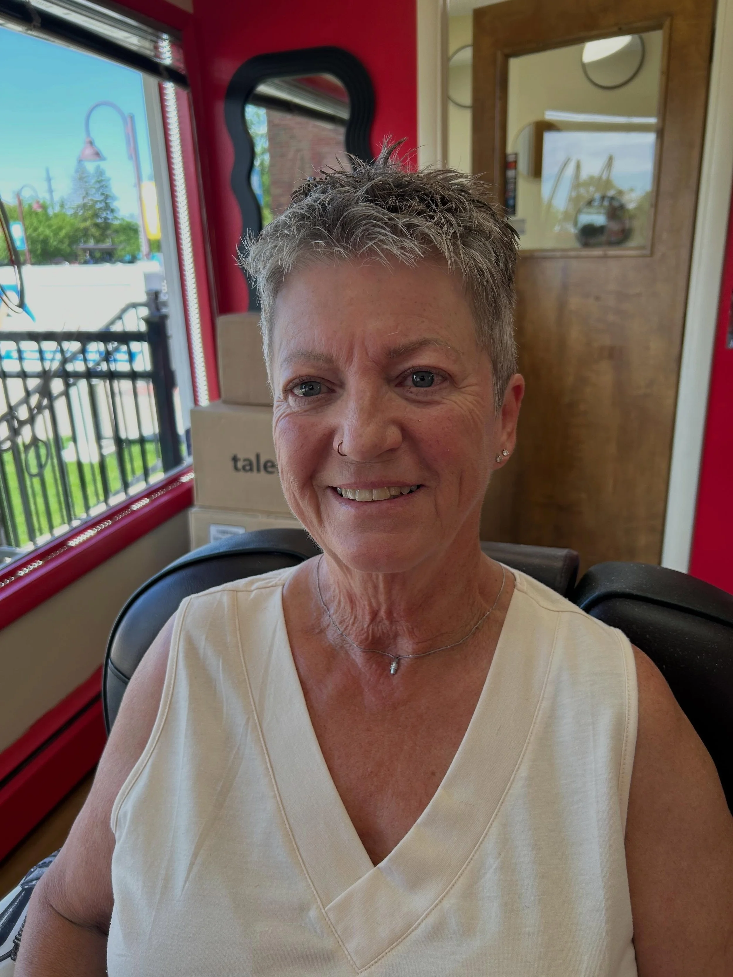 A smiling woman with short gray hair sitting inside a red and wood-paneled space, possibly a bus or train, with a window showing outdoor greenery and a sign that reads 'talent' in the background.