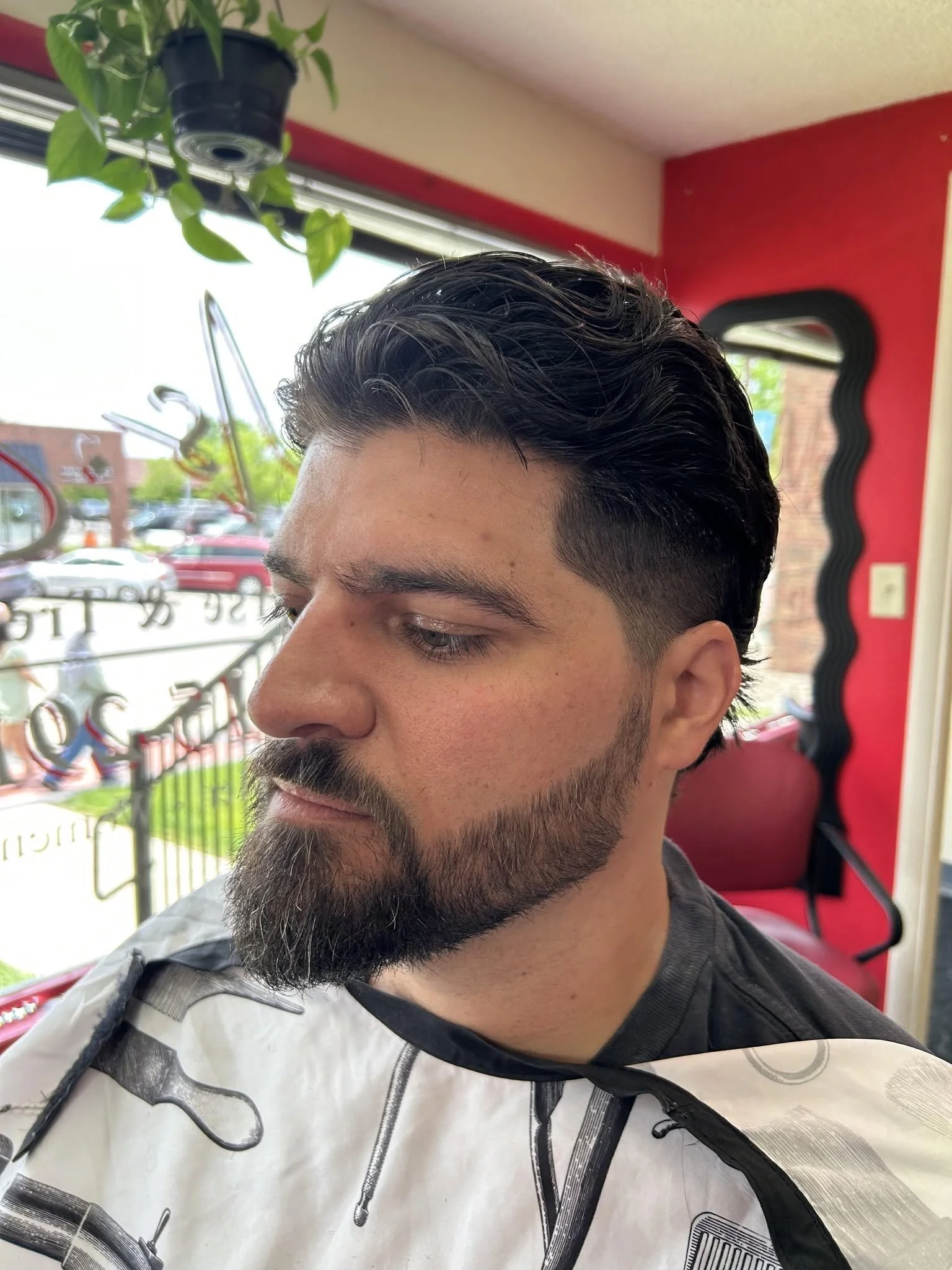 A man with dark, styled hair and a beard sitting inside a barber shop, wearing a barber cape with black and white patterns, with a red and black wall and a mirror in the background.
