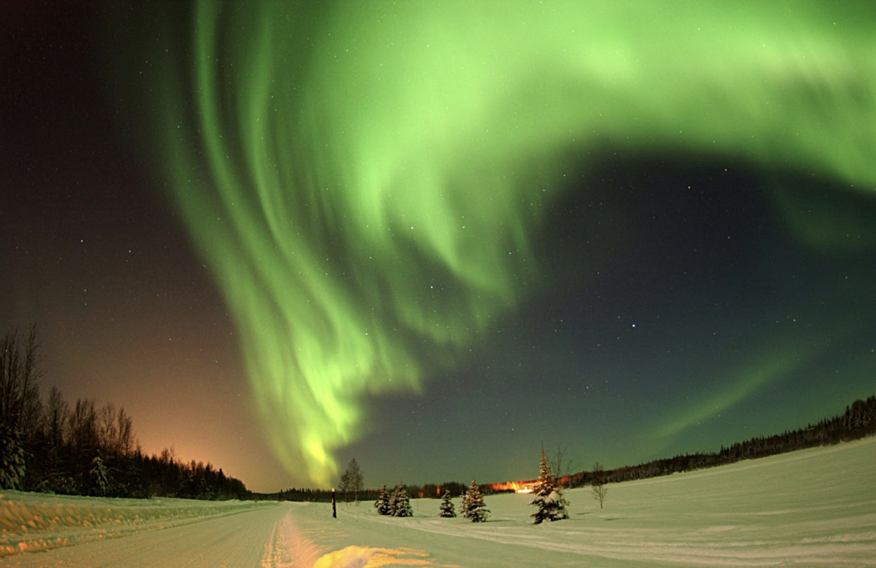 Northern lights (aurora borealis) over snowy landscape with trees and distant lights at night.