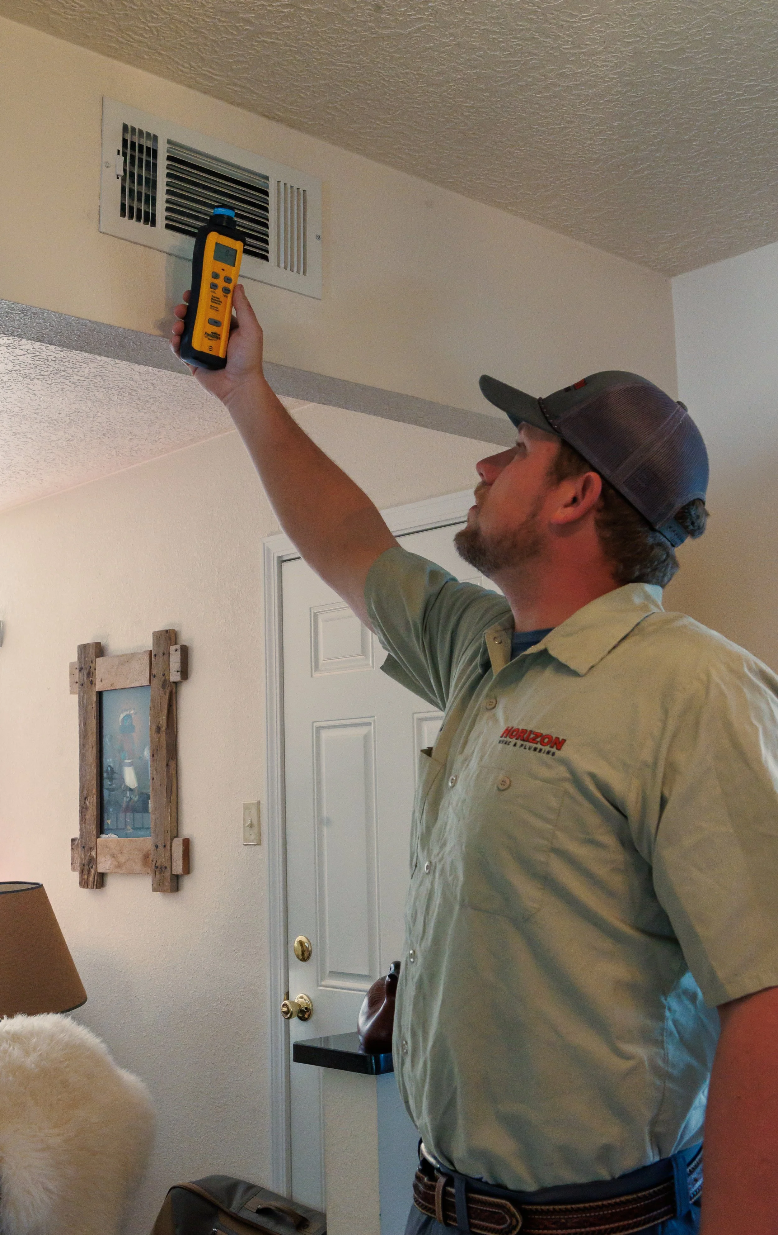 A man wearing a khaki work shirt and a cap is using a handheld infrared thermometer to measure temperature at an air vent on the ceiling of a room.