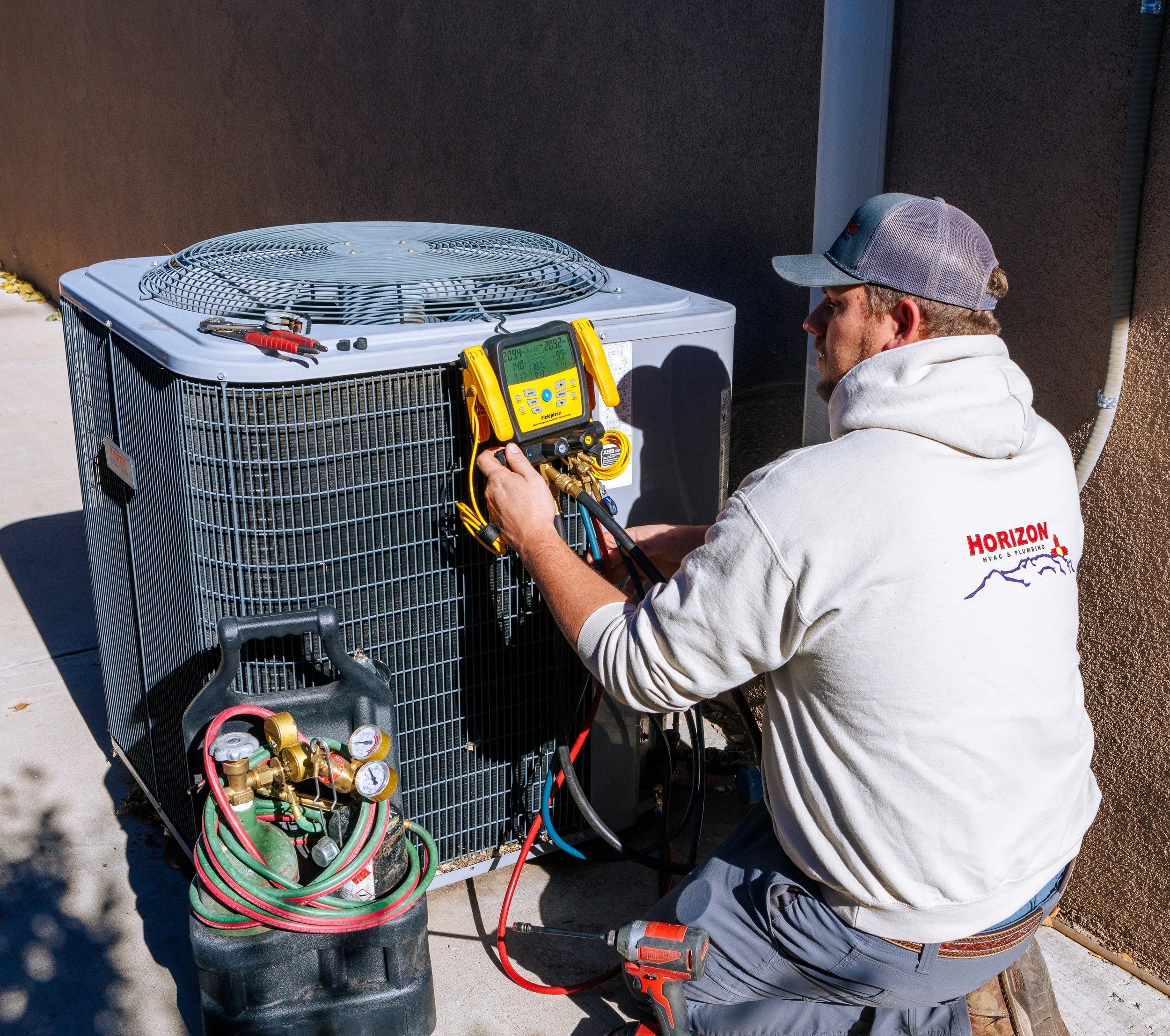 Technician working on a large outdoor air conditioning unit with diagnostic tools, wearing a hooded sweatshirt with the logo 'Horizon HVAC & Plumbing'.