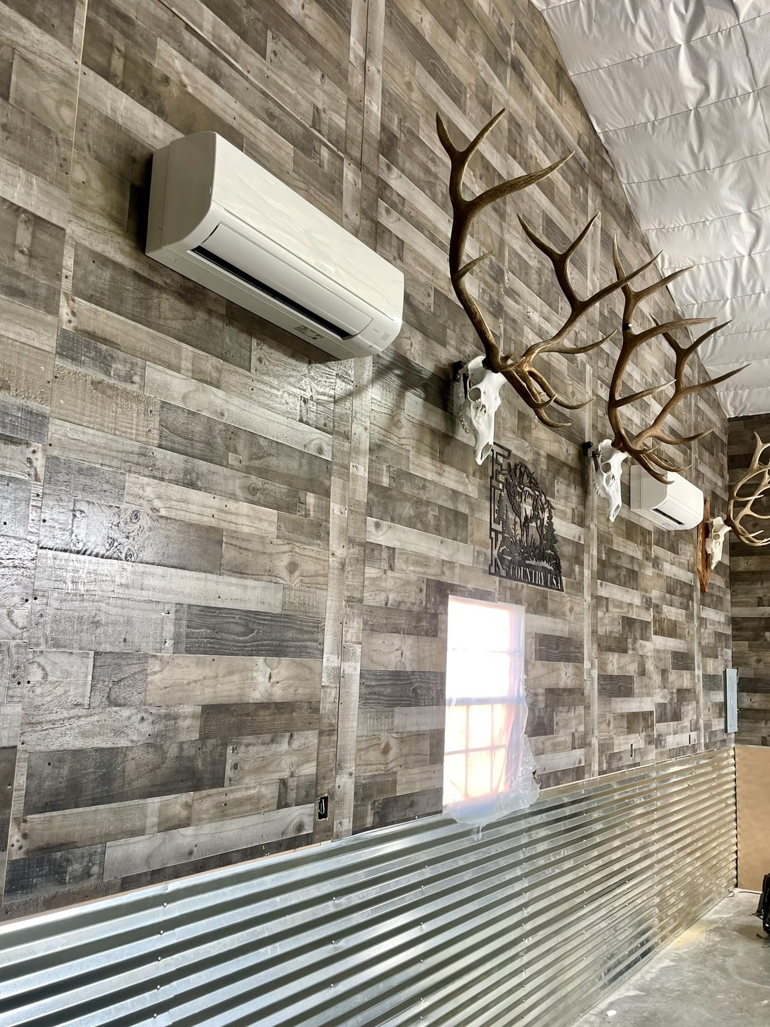 Wood-paneled wall with two mounted deer antlers, skulls, and air conditioning units. Small window with plastic covering and metal siding at the bottom.