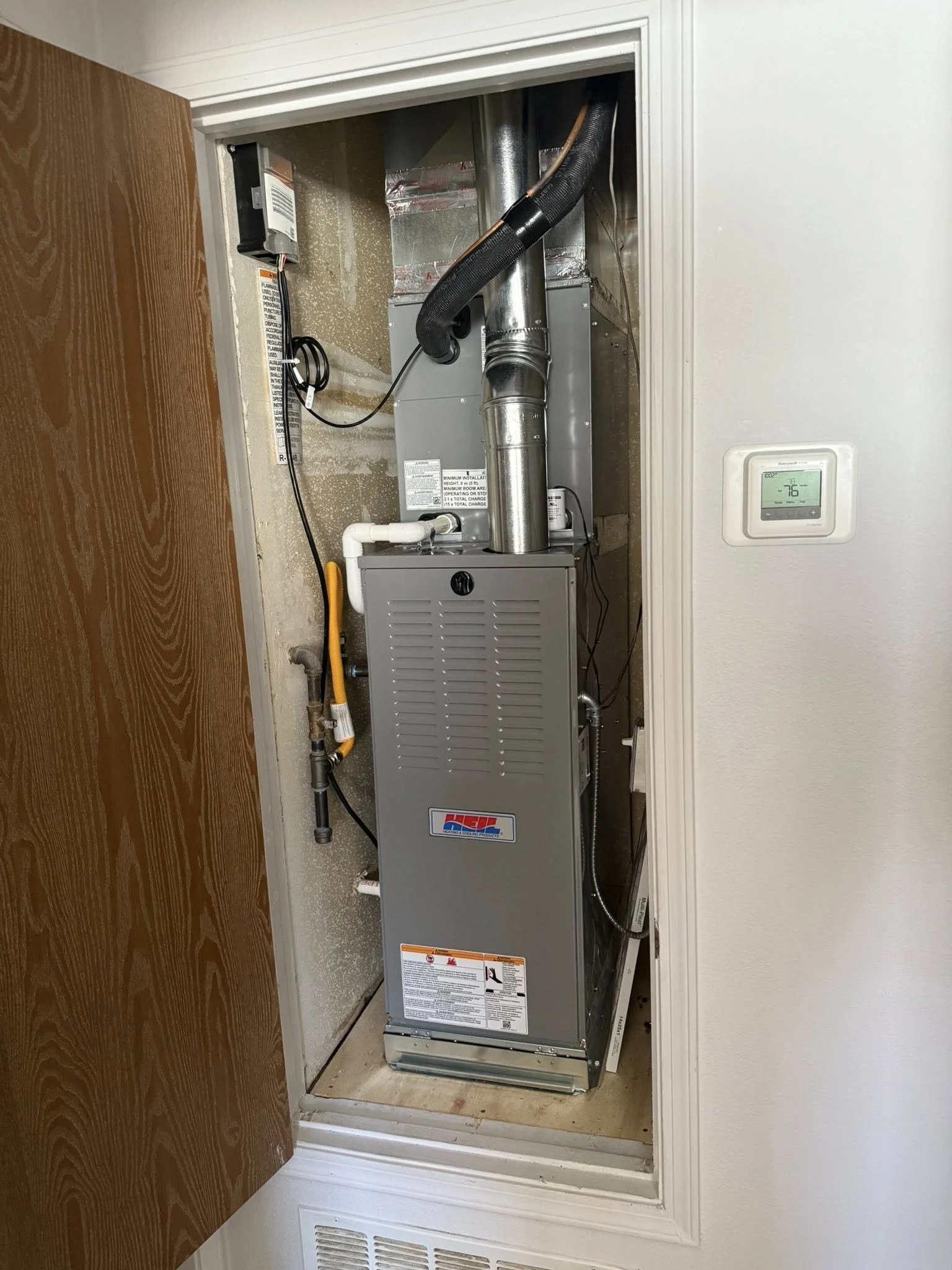 A furnace system installed inside a closet with a brown door open, featuring various pipes, ducts, and controls.
