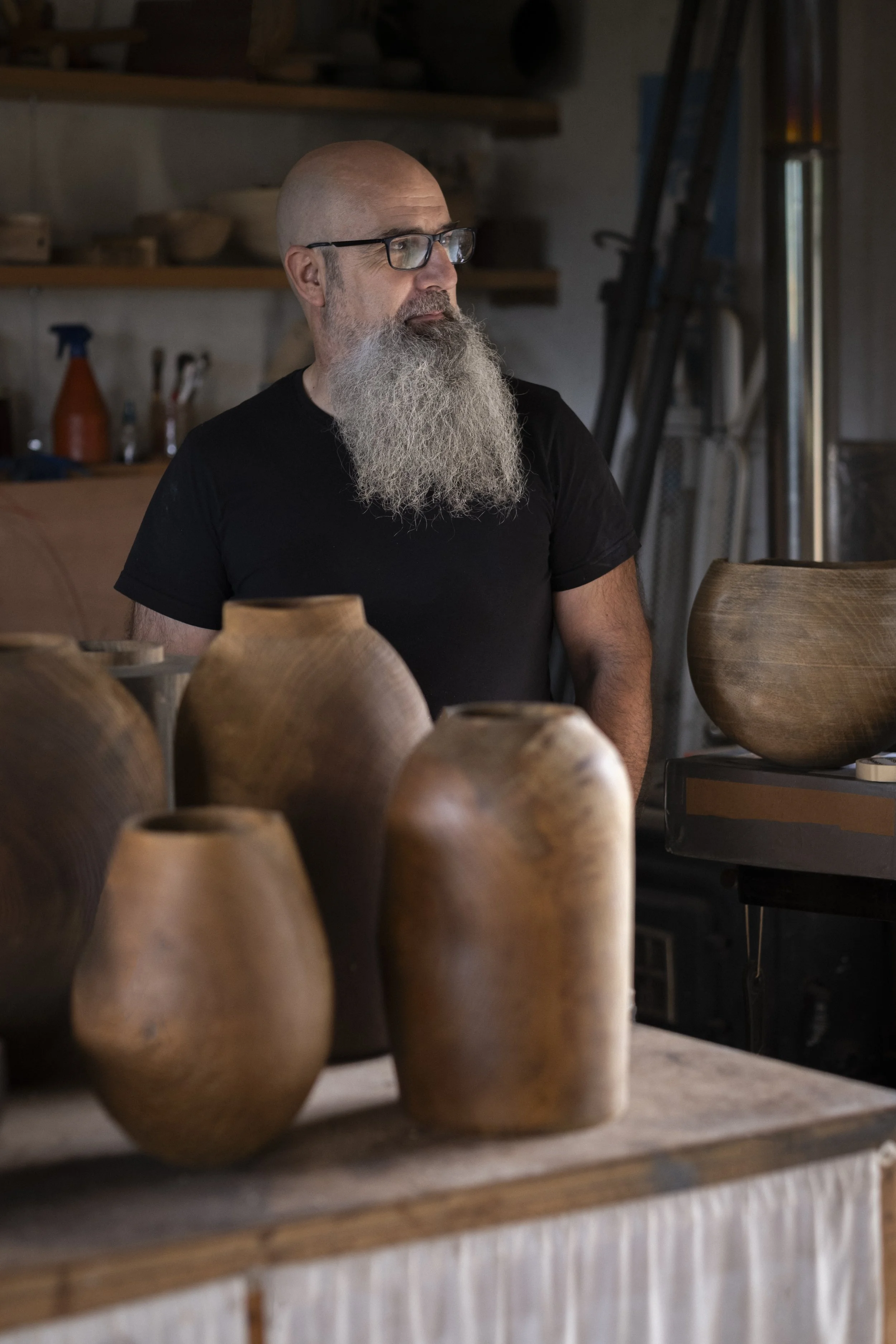 Hombre calvo de barba larga y gafas, vestido con camiseta negra, observando en un taller de cerámica rodeado de jarrones de barro.