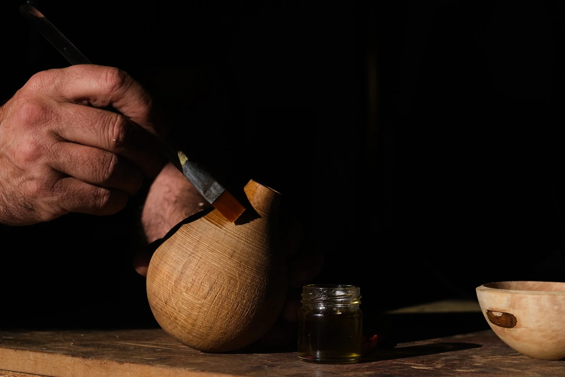 Mano tallando madera con herramienta de carpintería, en un ambiente oscuro.