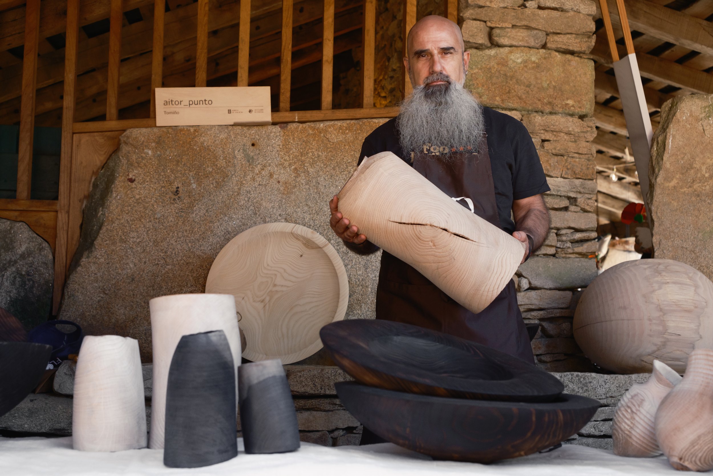 Hombre con barba sosteniendo una pieza de cerámica en una feria de artesanías, exhibiendo diferentes cerámicas y platos de madera y cerámica en una mesa.