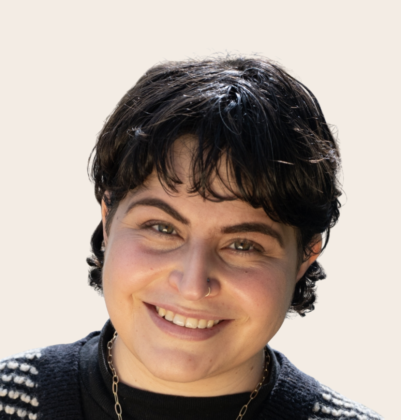 Close-up of a young woman with short black hair, smiling, wearing a black shirt with white stripes on the shoulders, a necklace, and a nose ring.