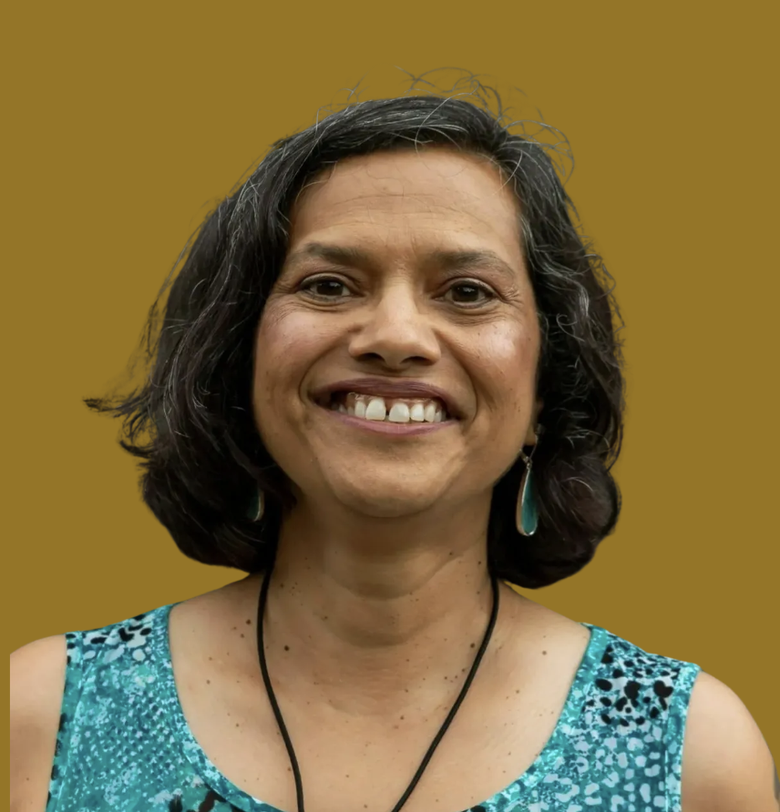 A woman smiling, with shoulder-length dark curly hair, wearing a turquoise patterned sleeveless top, turquoise earrings, and a black necklace, against a solid brown background.