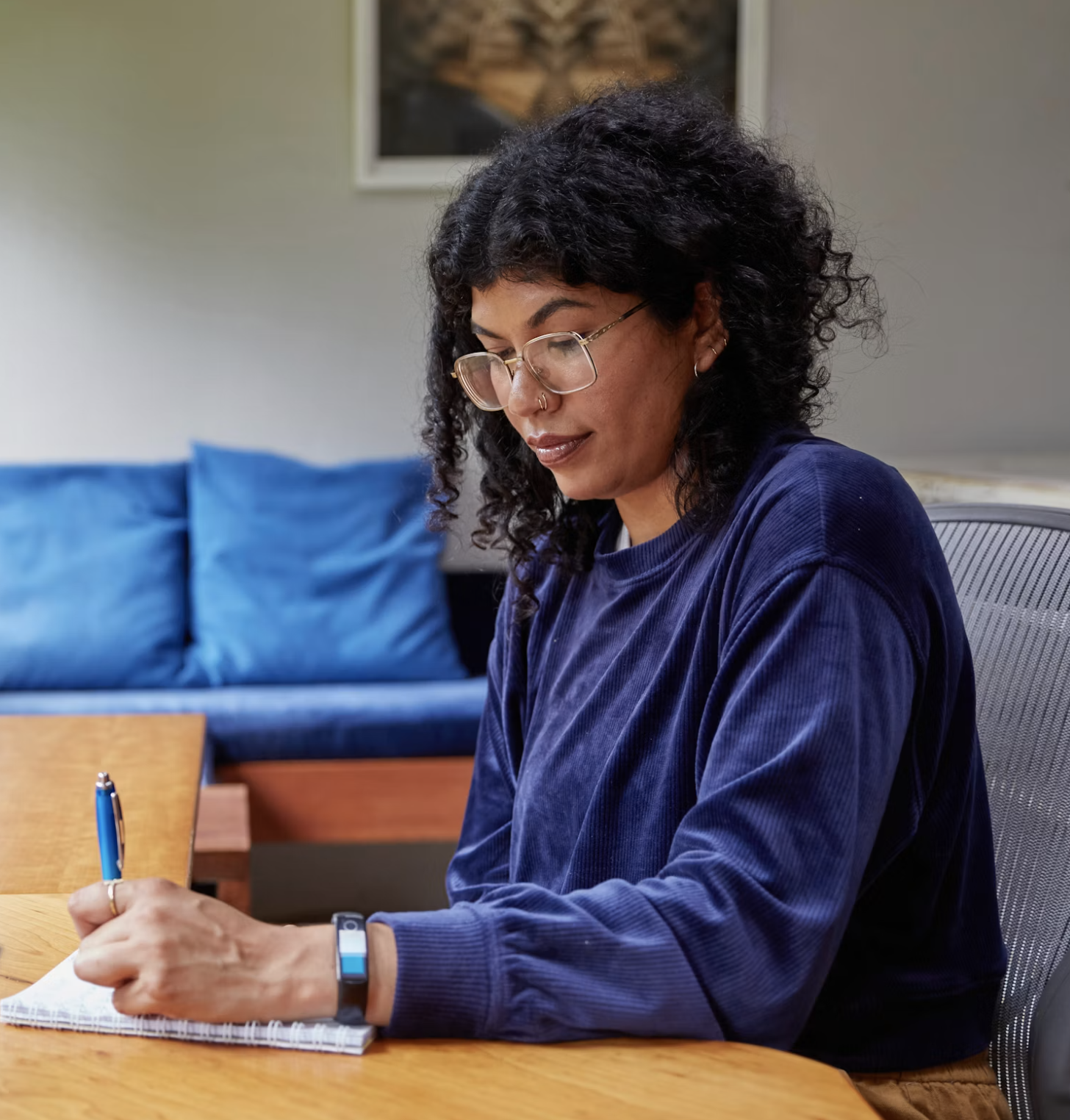 A woman with curly black hair, glasses, and earrings is sitting at a wooden table, writing in a notebook with a pen. She is wearing a blue velvety shirt and a fitness tracker on her left wrist. Behind her, a blue couch with pillows and a framed picture on the wall are visible.