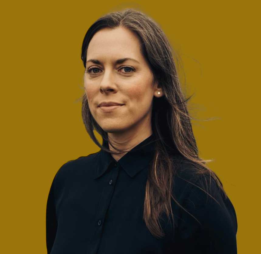 A woman with long brown hair wearing a black collared shirt, standing against a mustard yellow background.